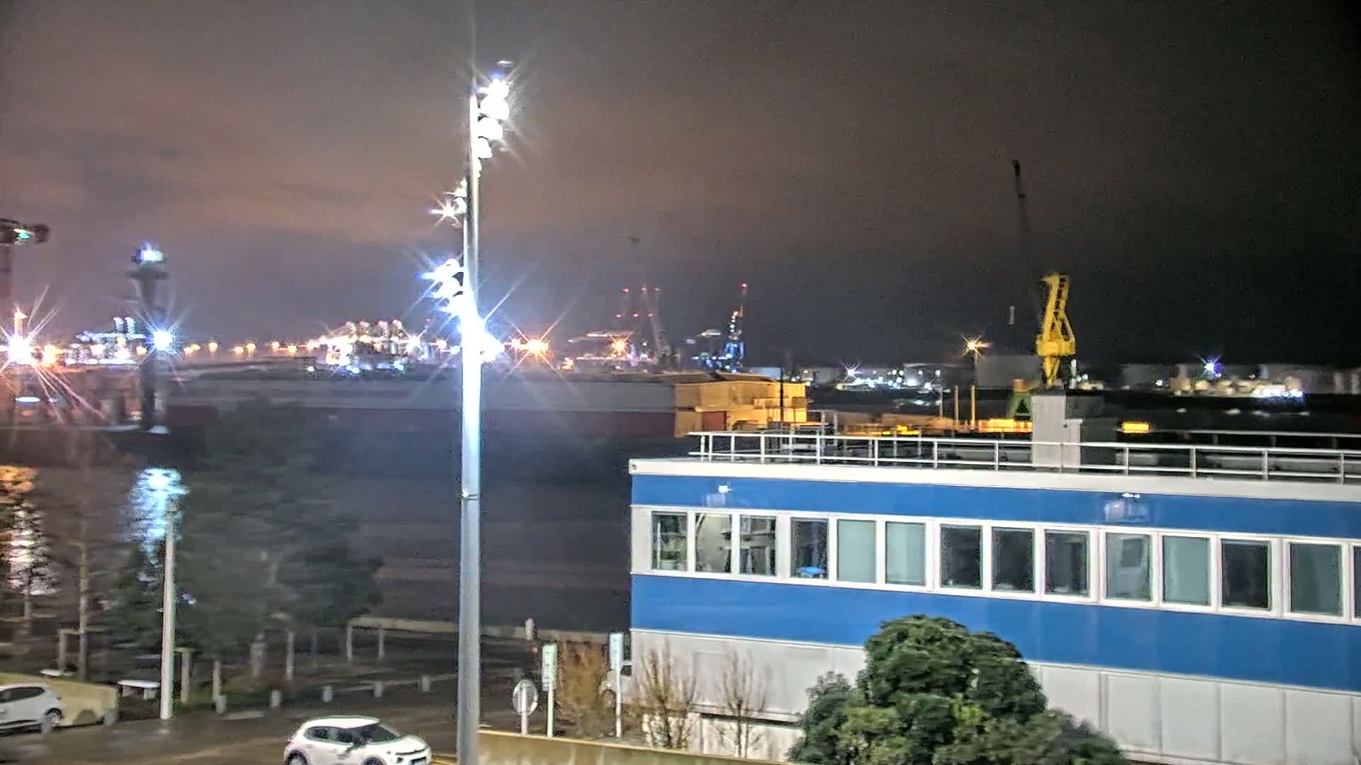 A nighttime view of a harbor with illuminated industrial buildings and cranes across a body of water, seen from a vantage point near a blue building and streetlights under a dark, cloudy sky.