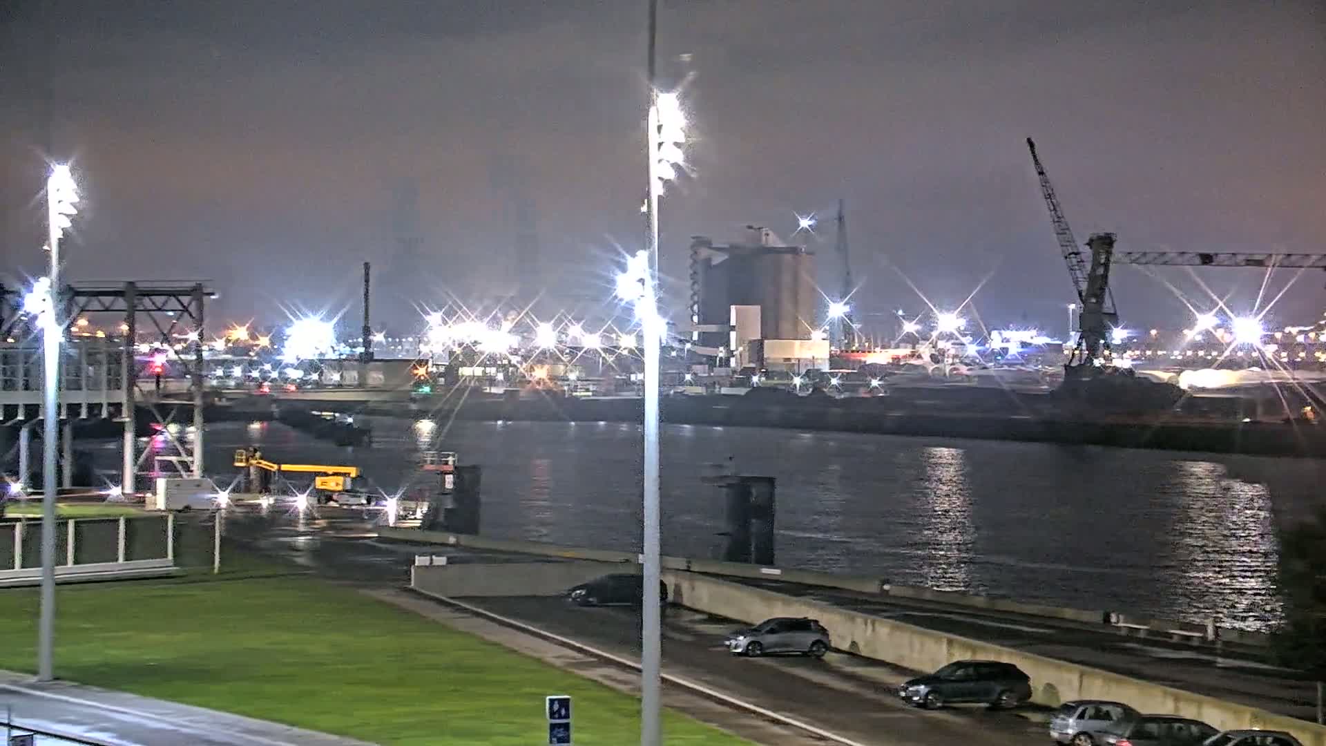 A nighttime view of a harbor with numerous lights illuminating the water and industrial structures, under a cloudy sky.