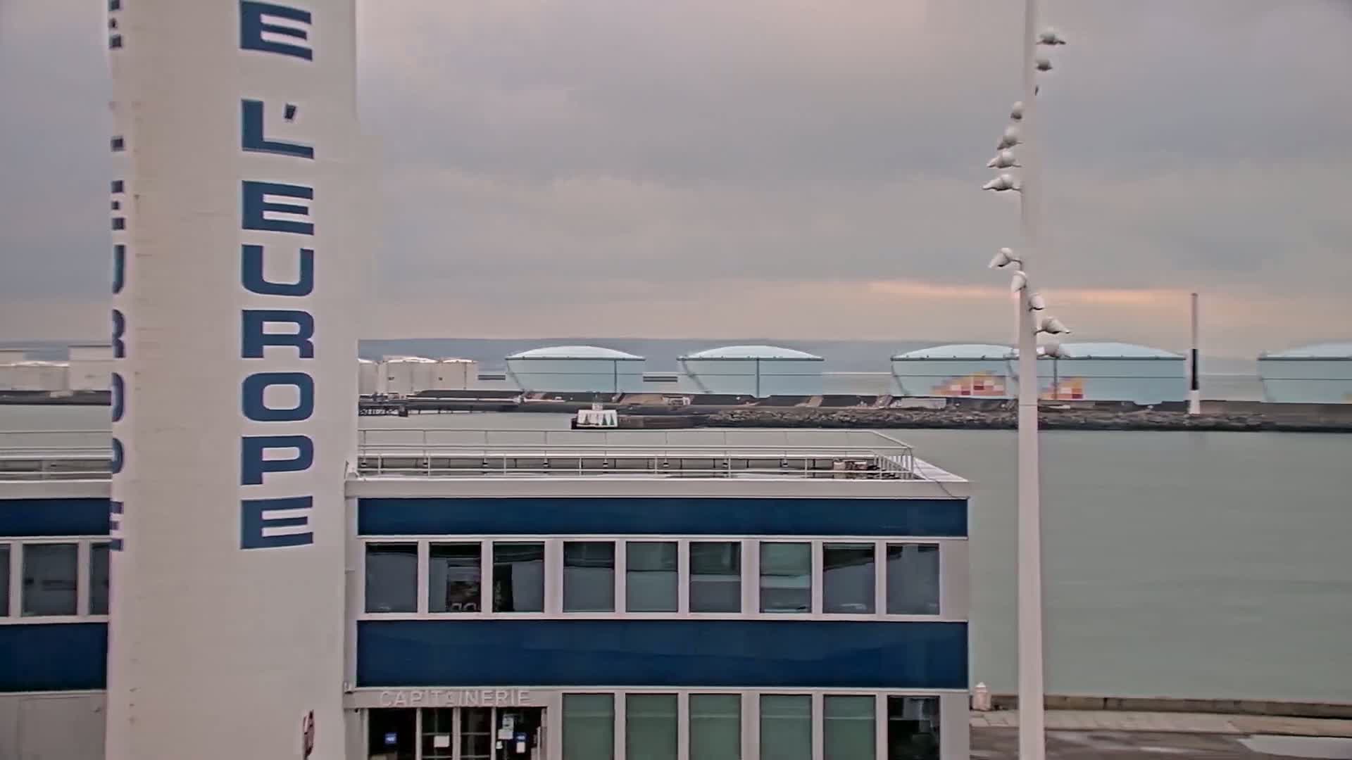 A blue building with a flat roof stands in front of a tall white tower with "Europe" written on it, overlooking a calm body of water with several large, light-blue storage tanks in the background under a cloudy sky.