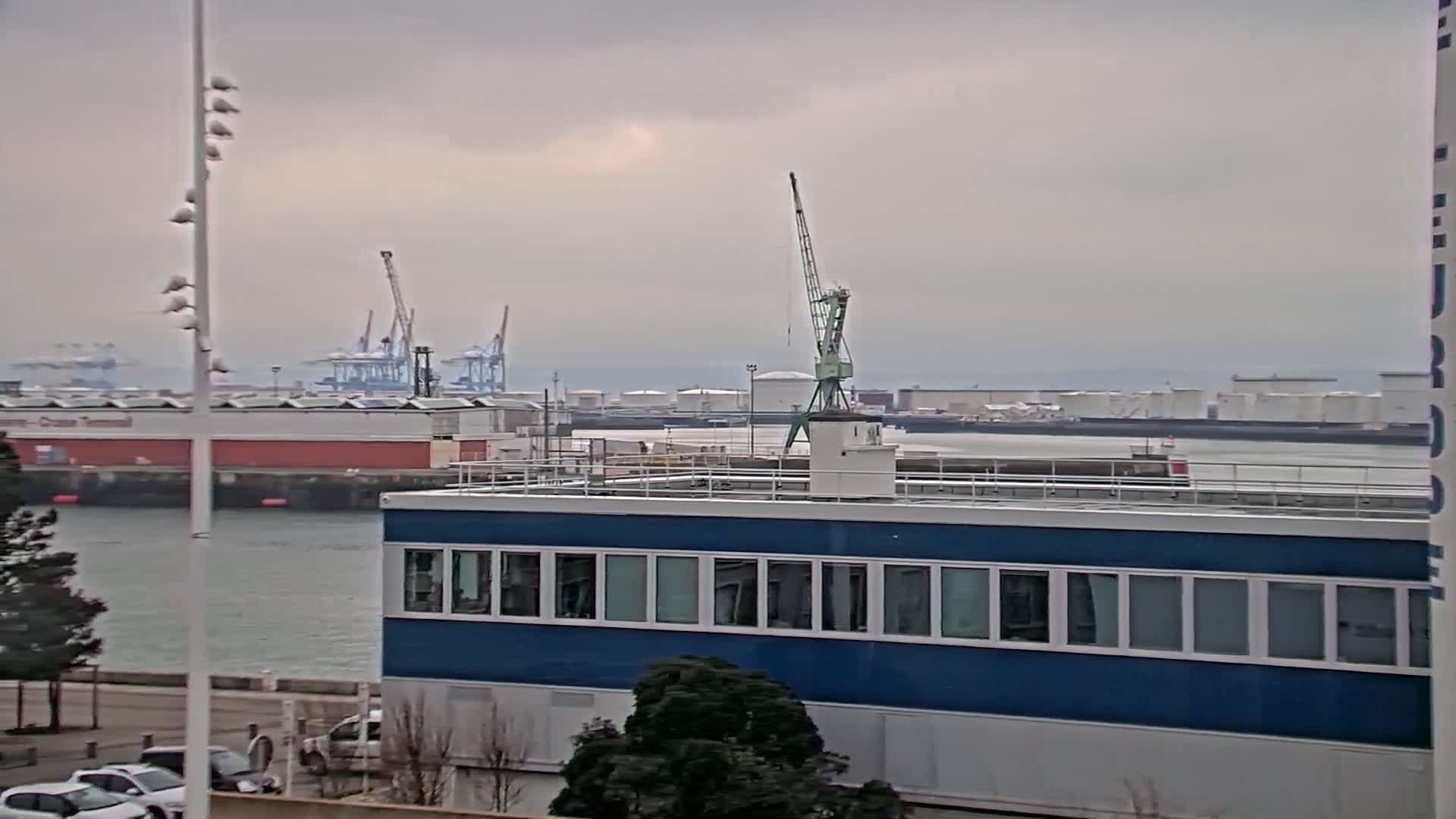 A blue building with many windows sits in the foreground overlooking a harbor with cranes, industrial buildings, and calm gray water under an overcast sky.