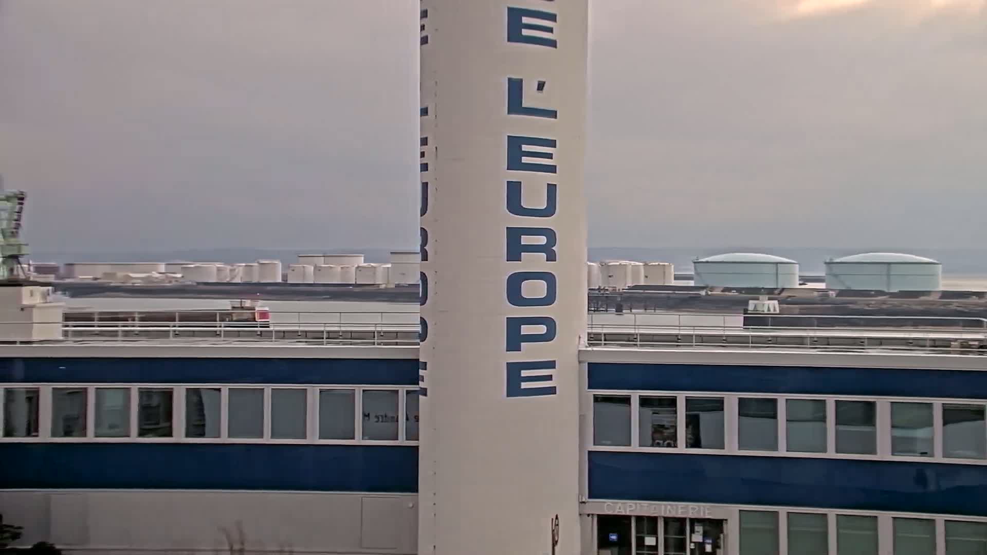 A tall white tower with "L'Europe" written on it stands in front of a blue building, overlooking a body of water with numerous storage tanks in the distance under an overcast sky.