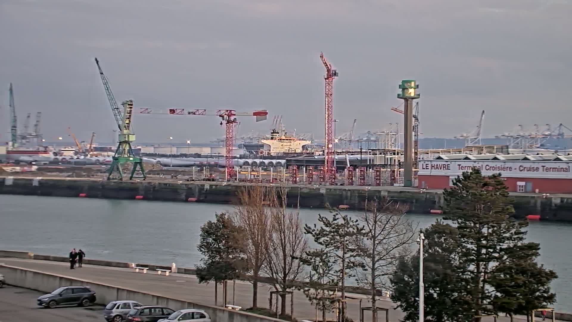 A harbor scene under an overcast sky shows several cranes, a ship, and a cruise terminal, with a waterfront walkway and bare trees in the foreground.
