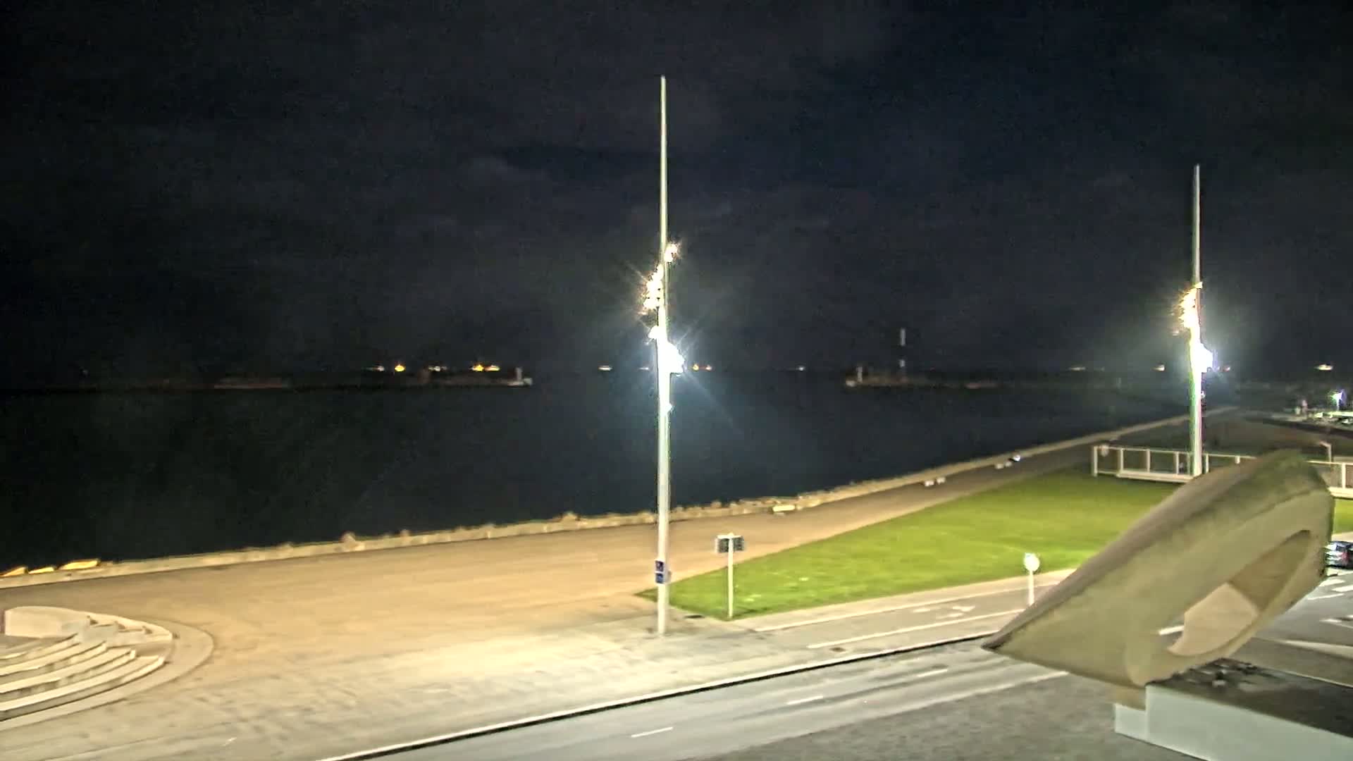 At night, a dark body of water is visible beyond a paved walkway and grassy area, lit by tall lampposts.