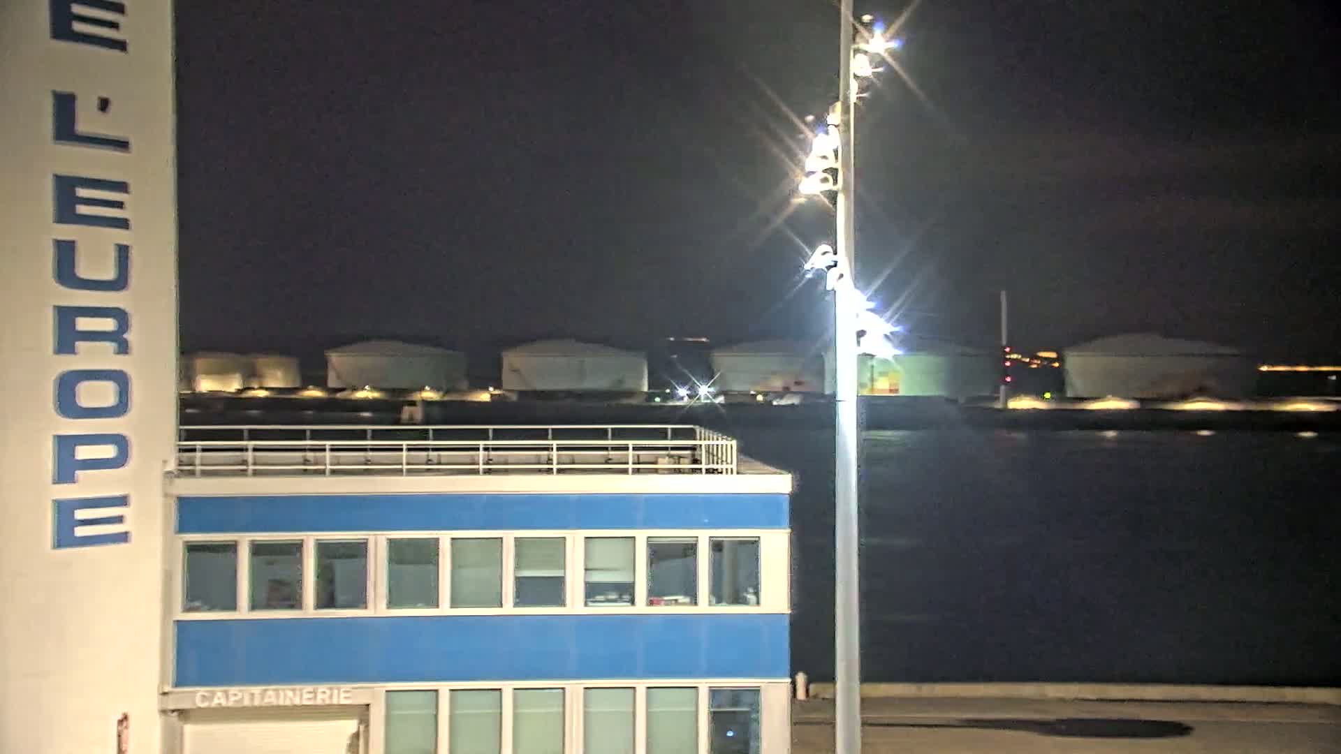 A brightly lit, blue building with a rooftop deck sits at night next to a tall light post, overlooking a dark body of water and several illuminated storage tanks in the distance.