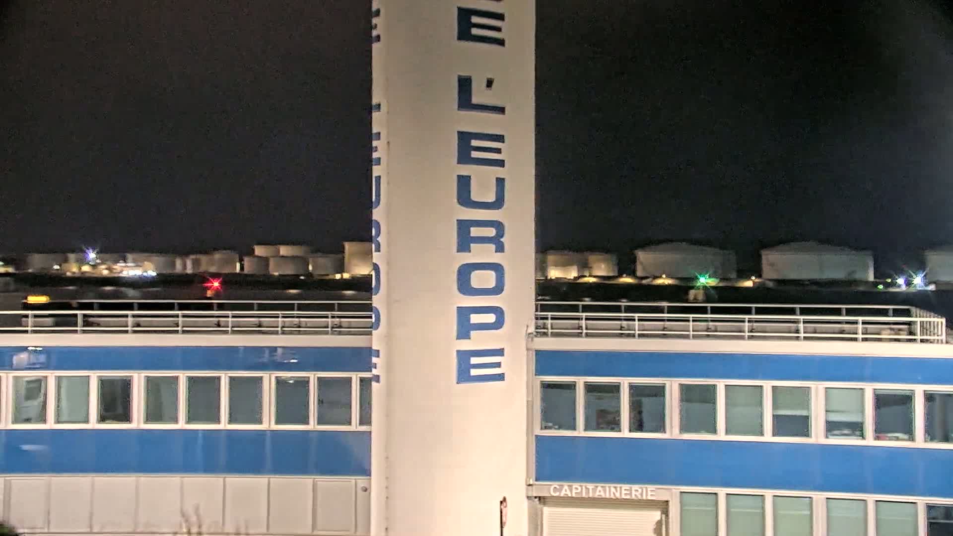 A tall, white tower with blue lettering stands in front of a blue building at night, with industrial storage tanks faintly visible in the background under a dark sky.