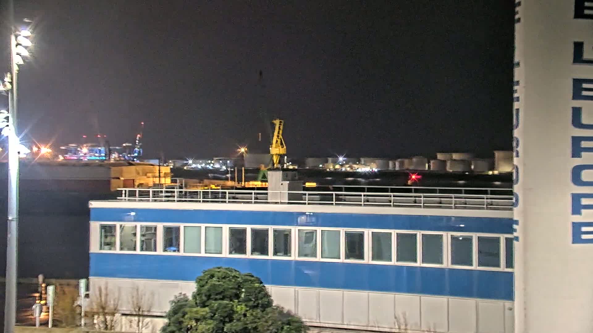 A nighttime view of a blue building with many windows, situated in front of an industrial port area with a crane and several lights visible in the distance.