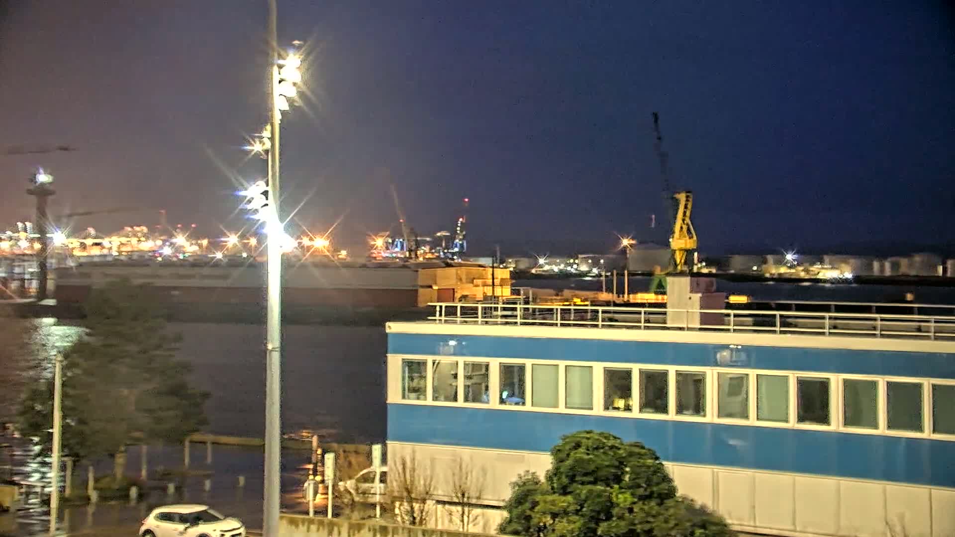 A nighttime view of a harbor with a blue building in the foreground, a body of water reflecting lights, and an industrial area with cranes and lights in the background.