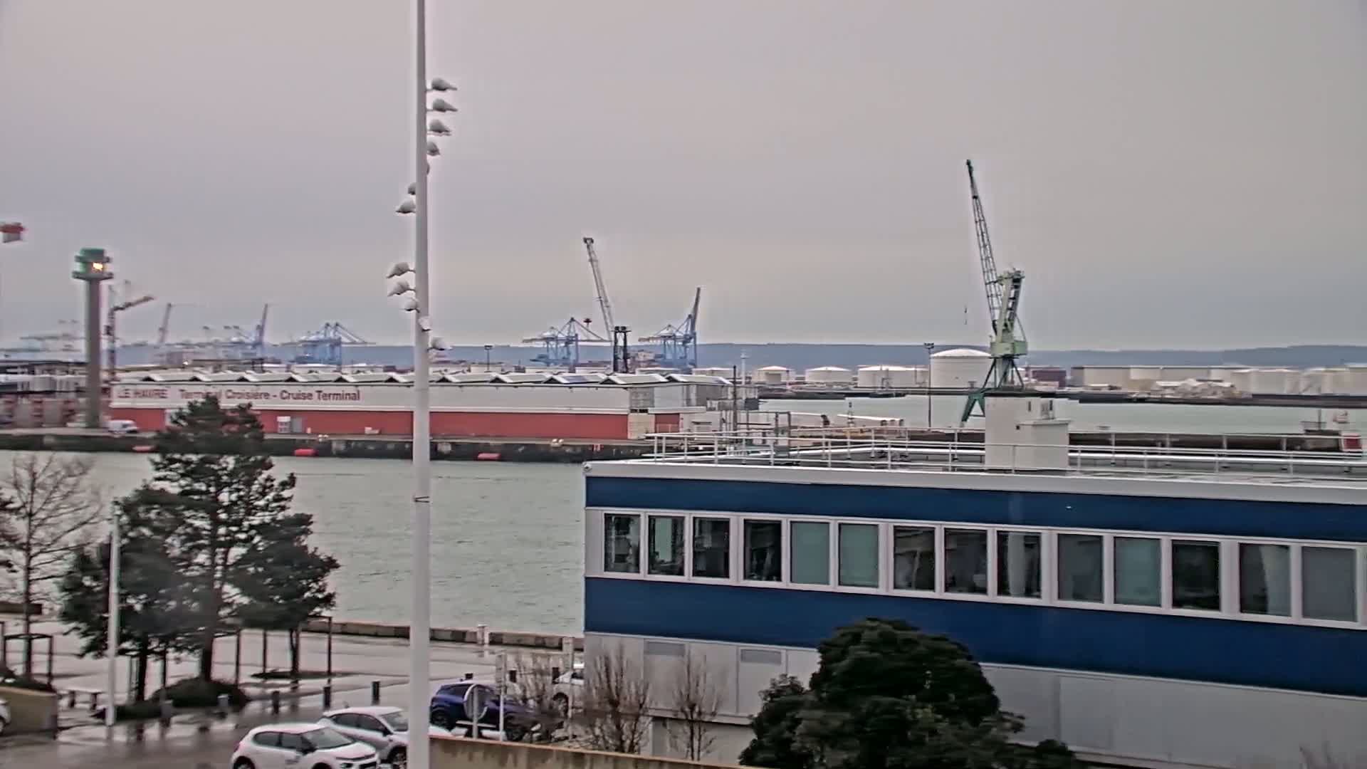 A harbor scene on an overcast day shows a blue building in the foreground, with a body of water, a cruise terminal, several cranes, and storage tanks in the background.