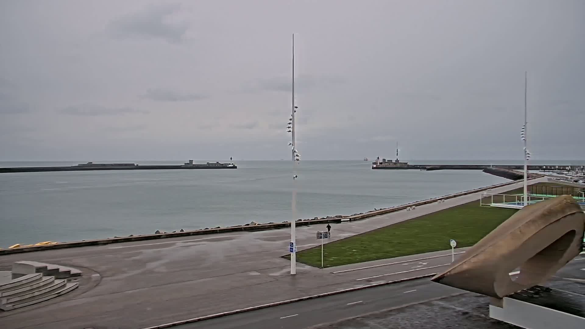 A coastal scene on an overcast day shows a harbor entrance with breakwaters, a grassy area, a road, and a large, abstract sculpture.