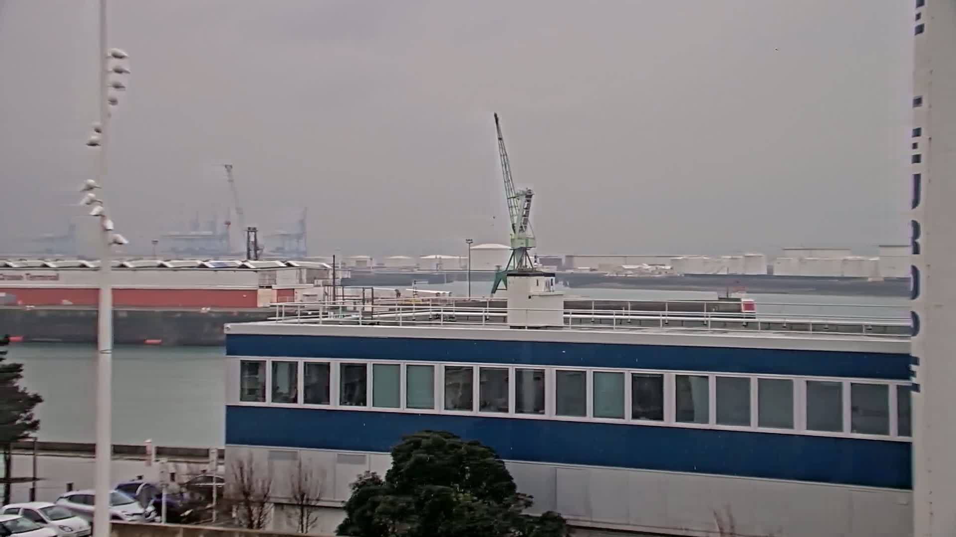 A blue building with many windows sits on a waterfront, a crane is visible on its roof, and a hazy gray sky indicates overcast weather.