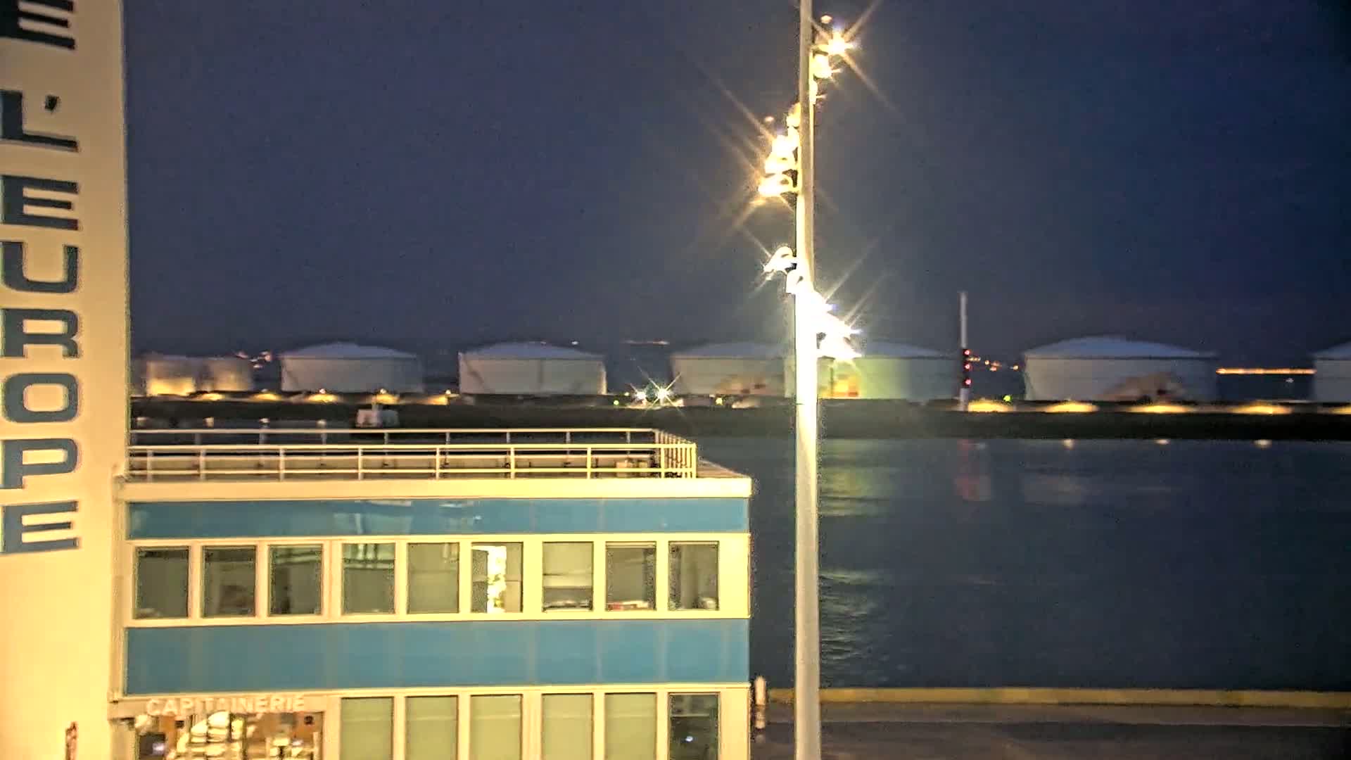 At night, a brightly lit building with multiple windows overlooks a body of dark water where several large storage tanks are visible in the distance.