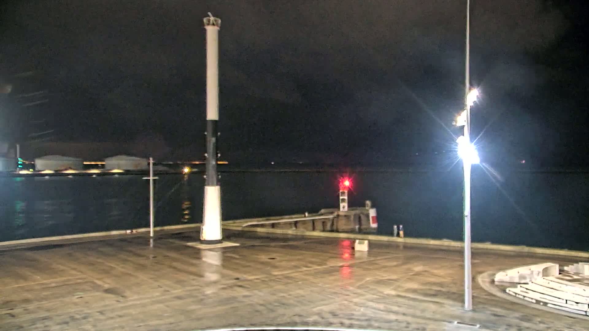 A tall, black and white lighthouse stands on a wet, dark harbor at night, with a smaller red light visible across the water.