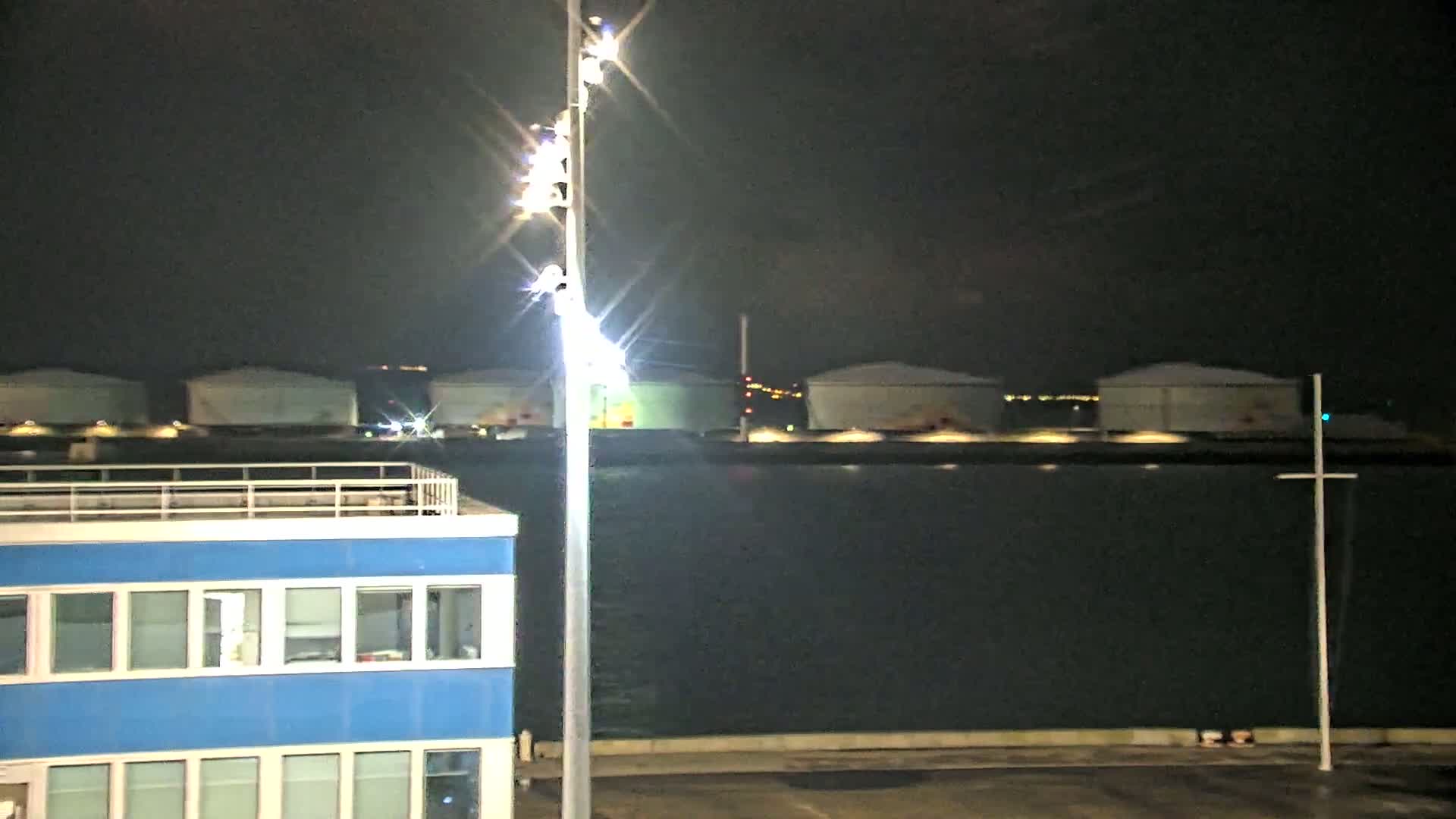 At night, a brightly lit tall pole stands near a blue building overlooking a dark body of water beyond which several large, light-colored structures are visible.