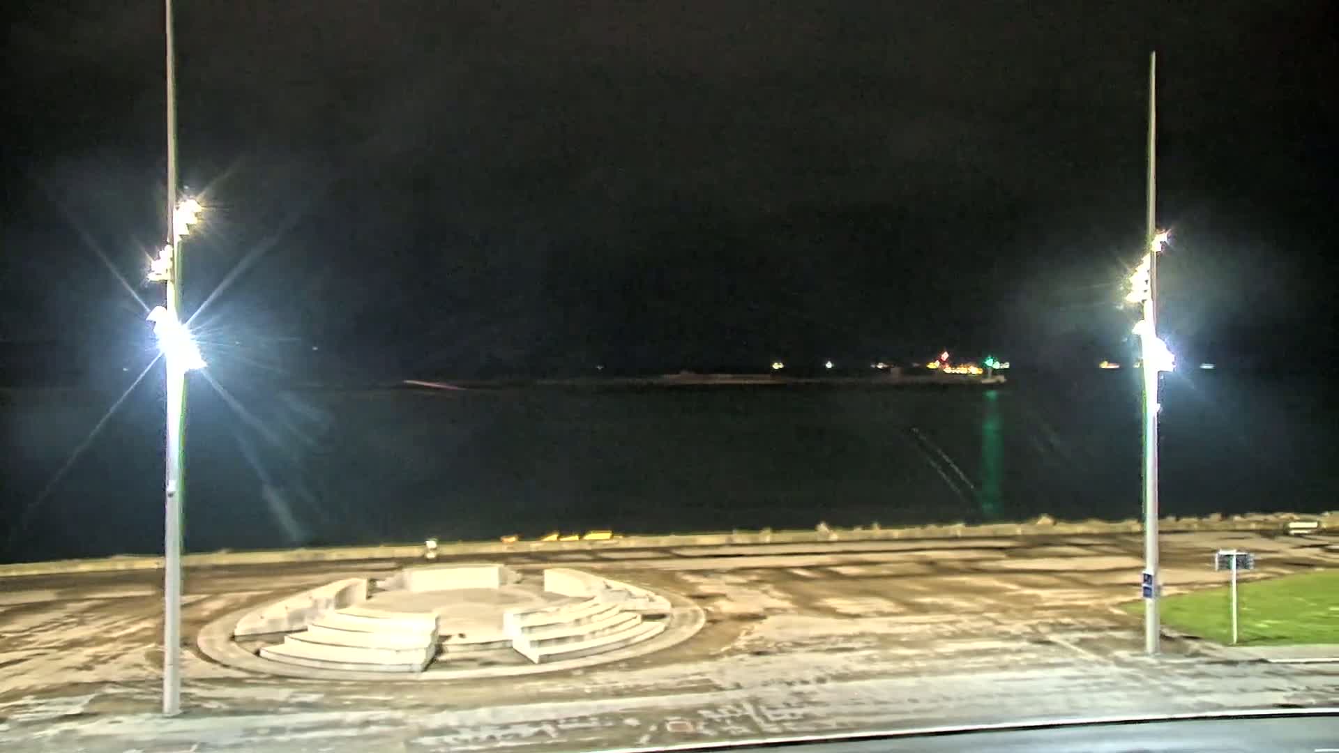 A circular stone structure sits in a mostly empty paved area at night next to a dark body of water under brightly lit tall lampposts; some snow or ice is visible on the ground.