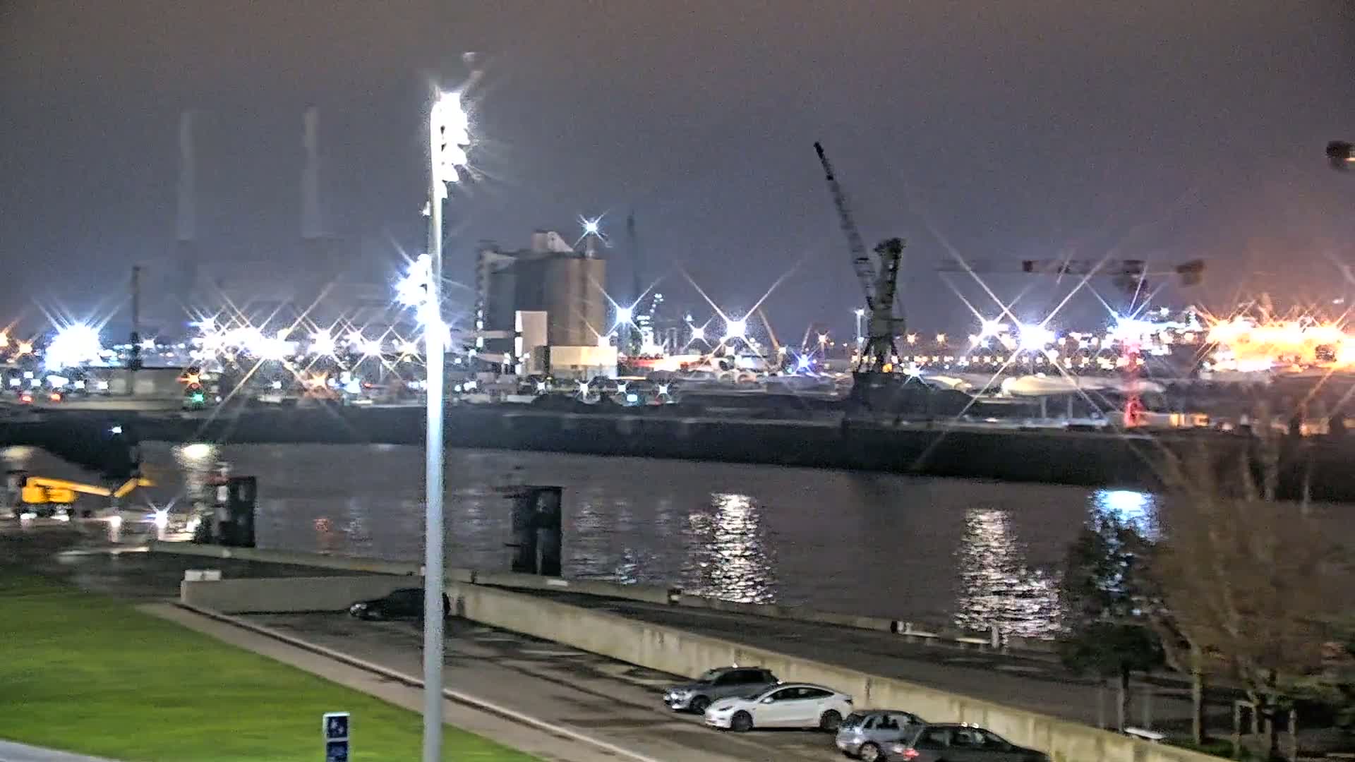 A nighttime view of a harbor with brightly lit industrial buildings and docked ships, reflecting in calm water.