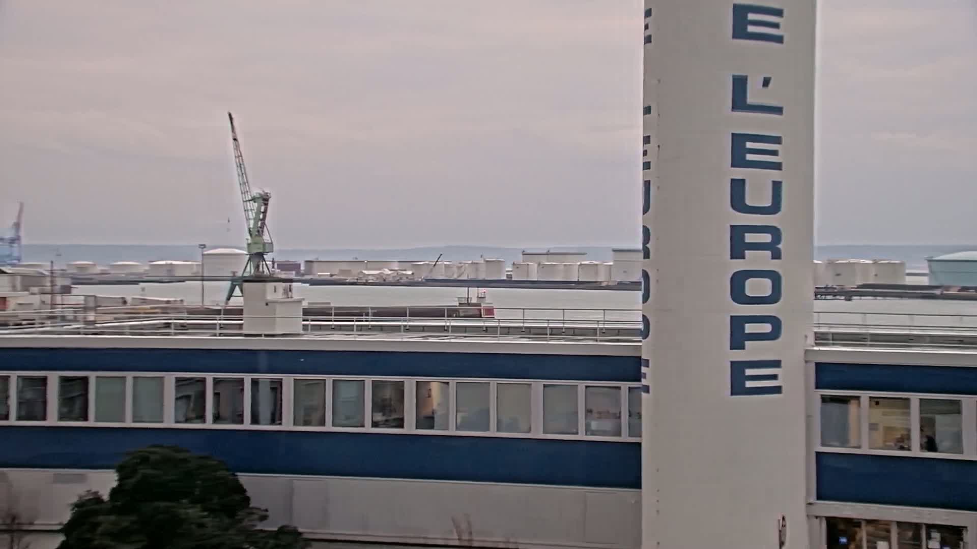 A view of an industrial area on an overcast day shows a building with a blue facade, a crane, and numerous storage tanks across a body of water in the distance.