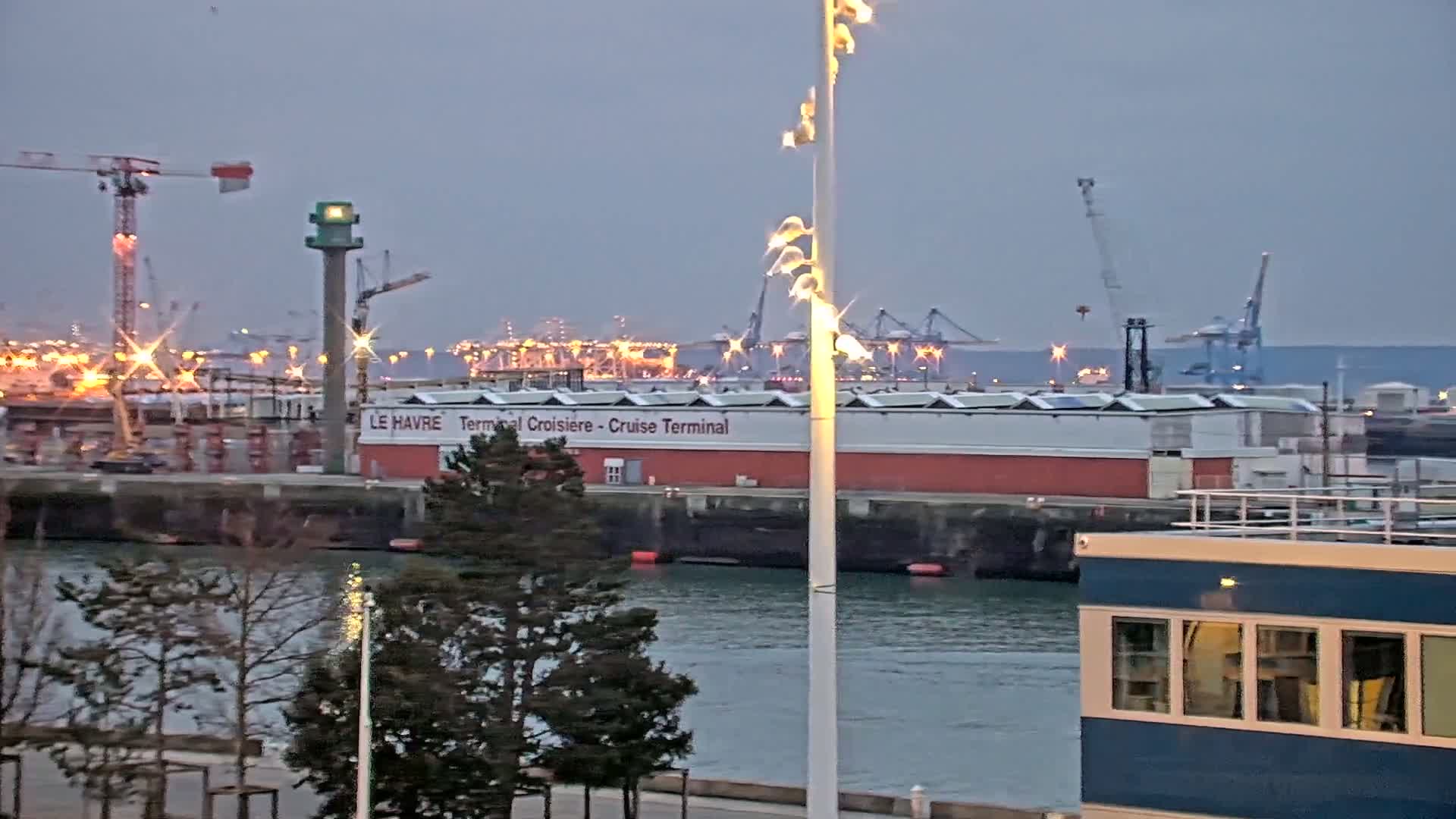 A harbor at dusk shows a cruise terminal, several cranes, and other industrial buildings, with calm water in the foreground and trees along a waterfront walkway.