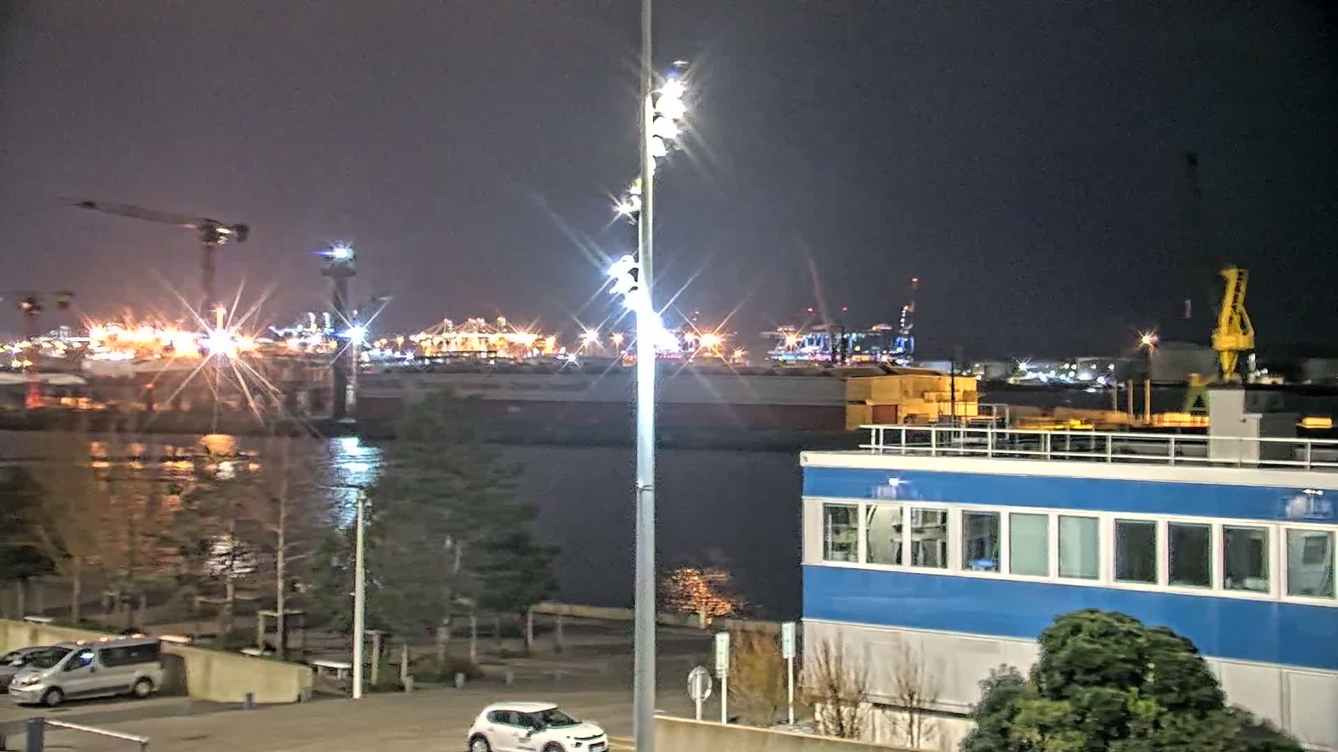 A nighttime view of a harbor with brightly lit industrial buildings and cranes across the water, a blue building in the foreground, and cars parked in a lot.