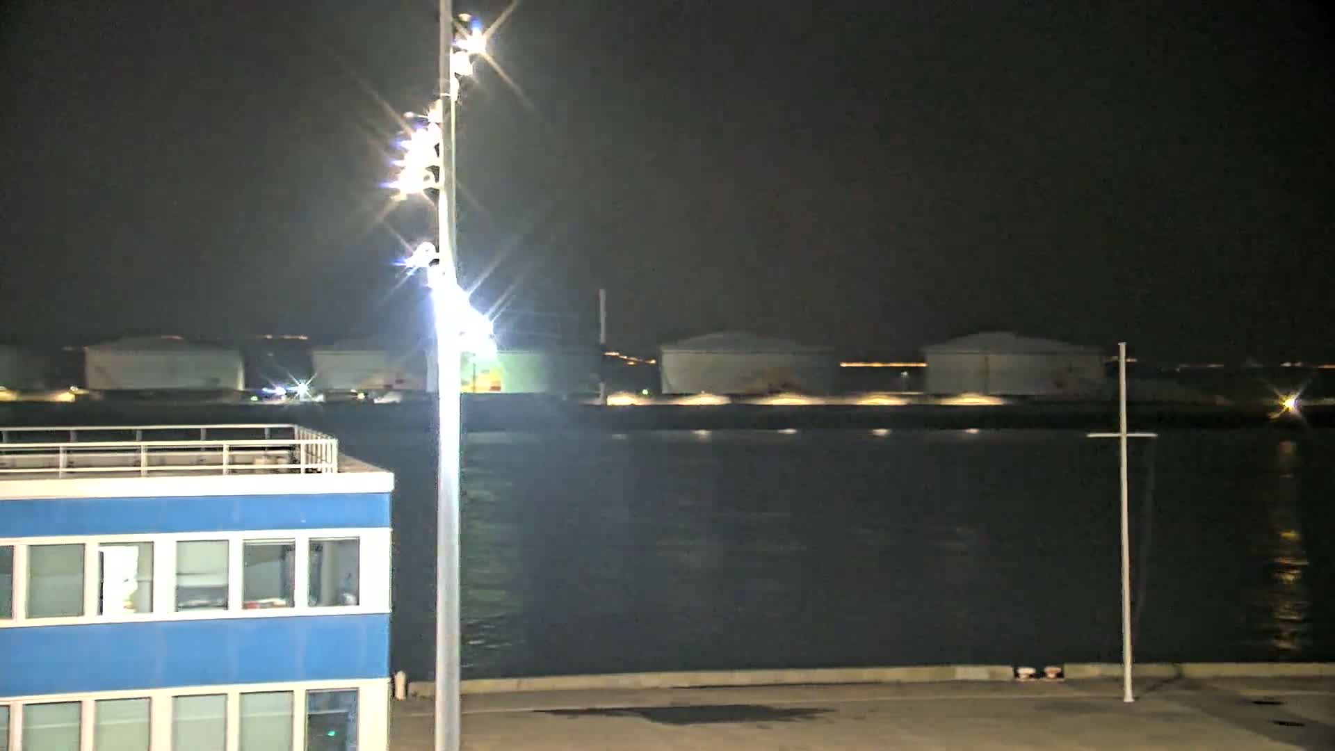 A brightly lit, blue building is situated near a body of dark water at night, with several large storage tanks visible across the water.