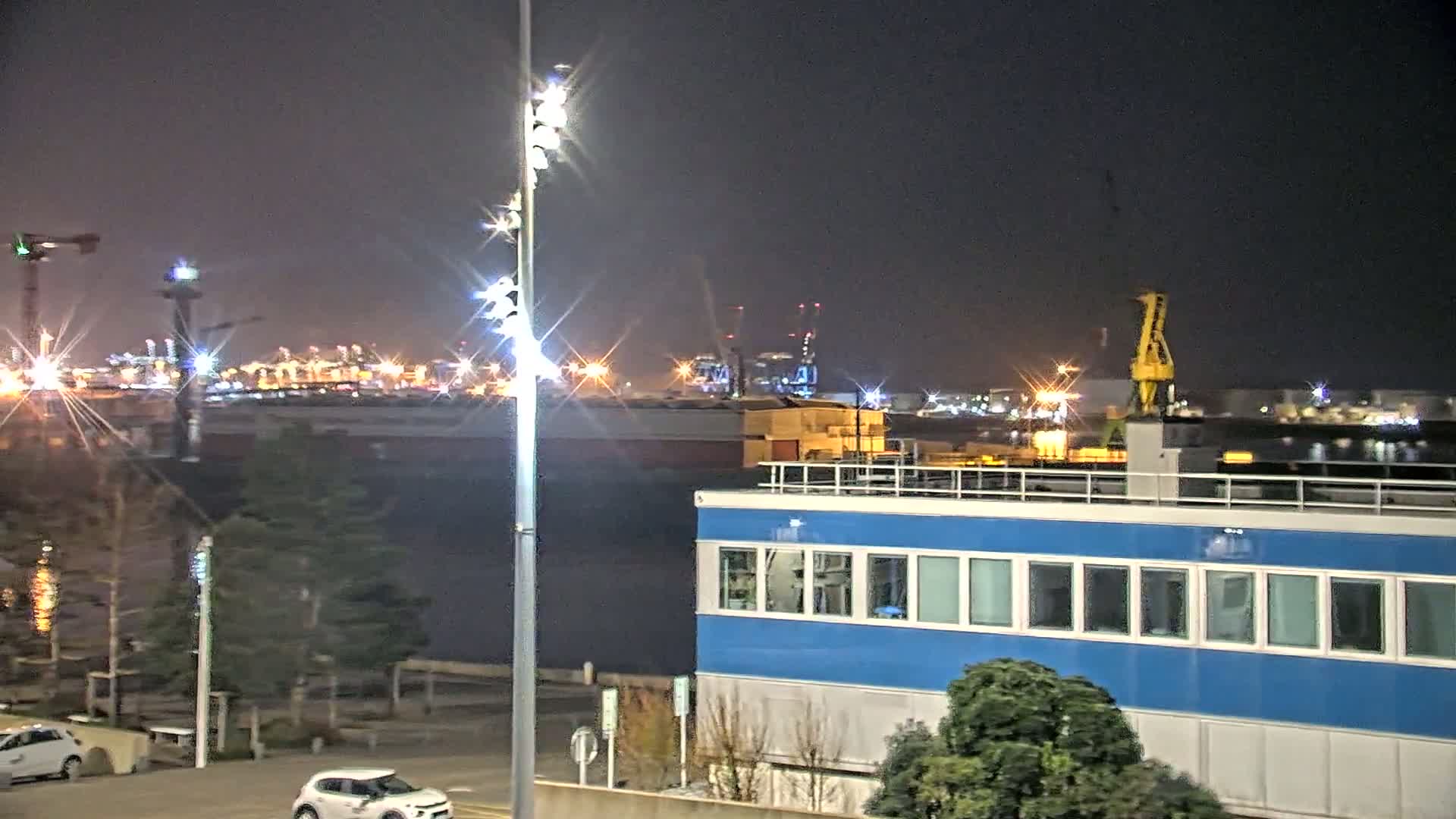 A nighttime view of a harbor with a brightly lit industrial area in the background, a blue building in the foreground, and a few cars parked nearby.