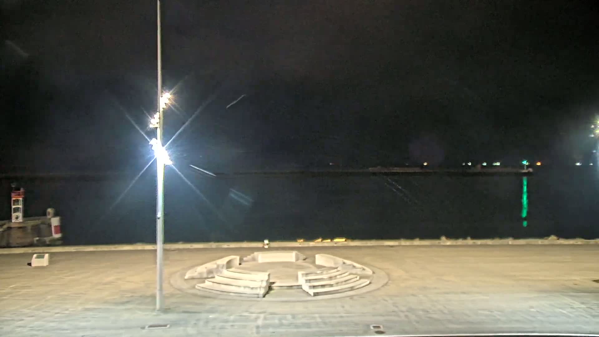 A circular stone seating area is situated on a waterfront plaza at night, with brightly lit lampposts and a calm dark body of water visible in the background.