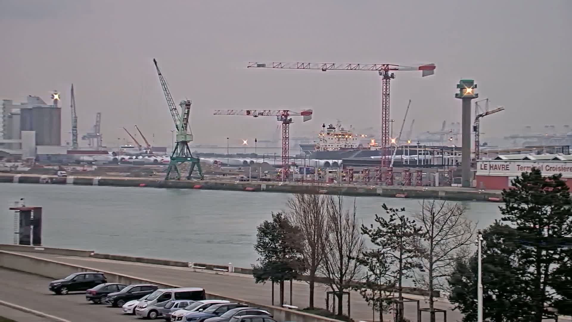 A harbor with several cranes, a large ship, and other industrial buildings is seen on an overcast day, with cars parked along a waterfront road in the foreground.