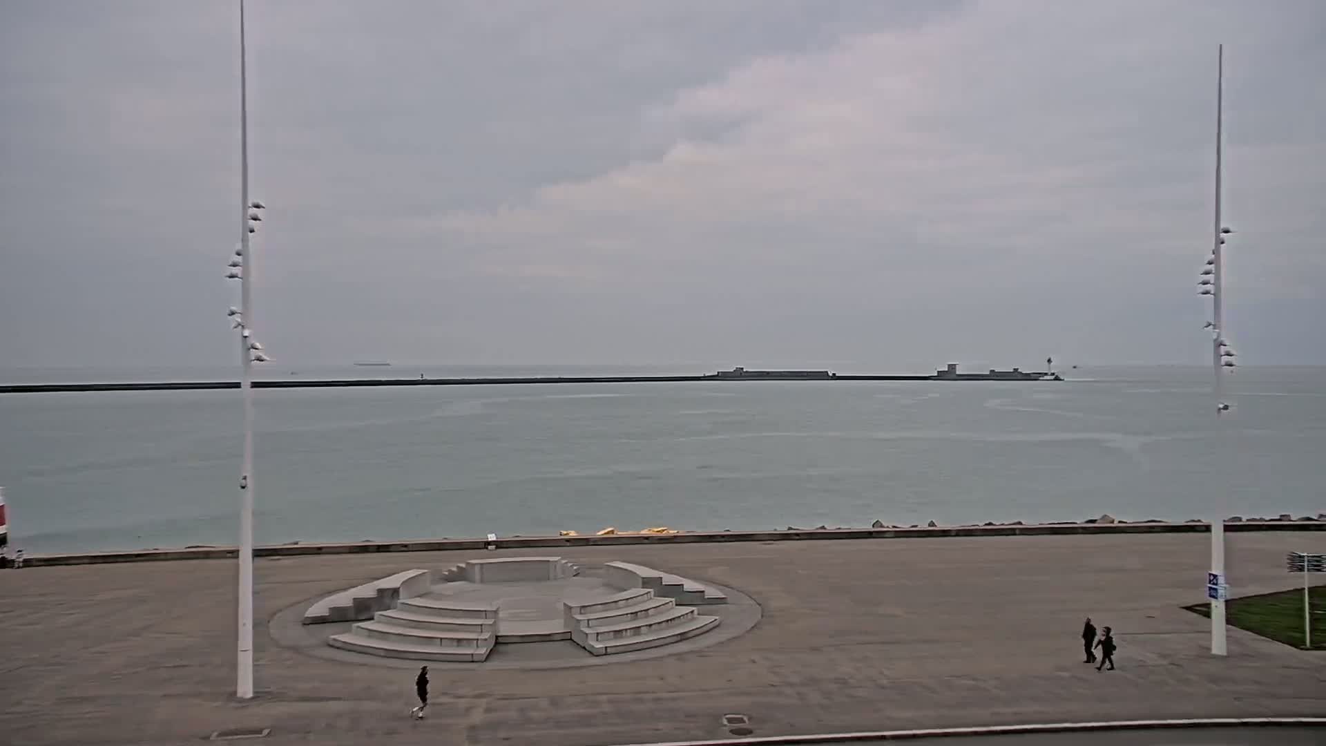A gray, overcast day reveals a seaside plaza with a low, circular stone structure, two tall light poles, and a few pedestrians near the water's edge, a breakwater in the distance.