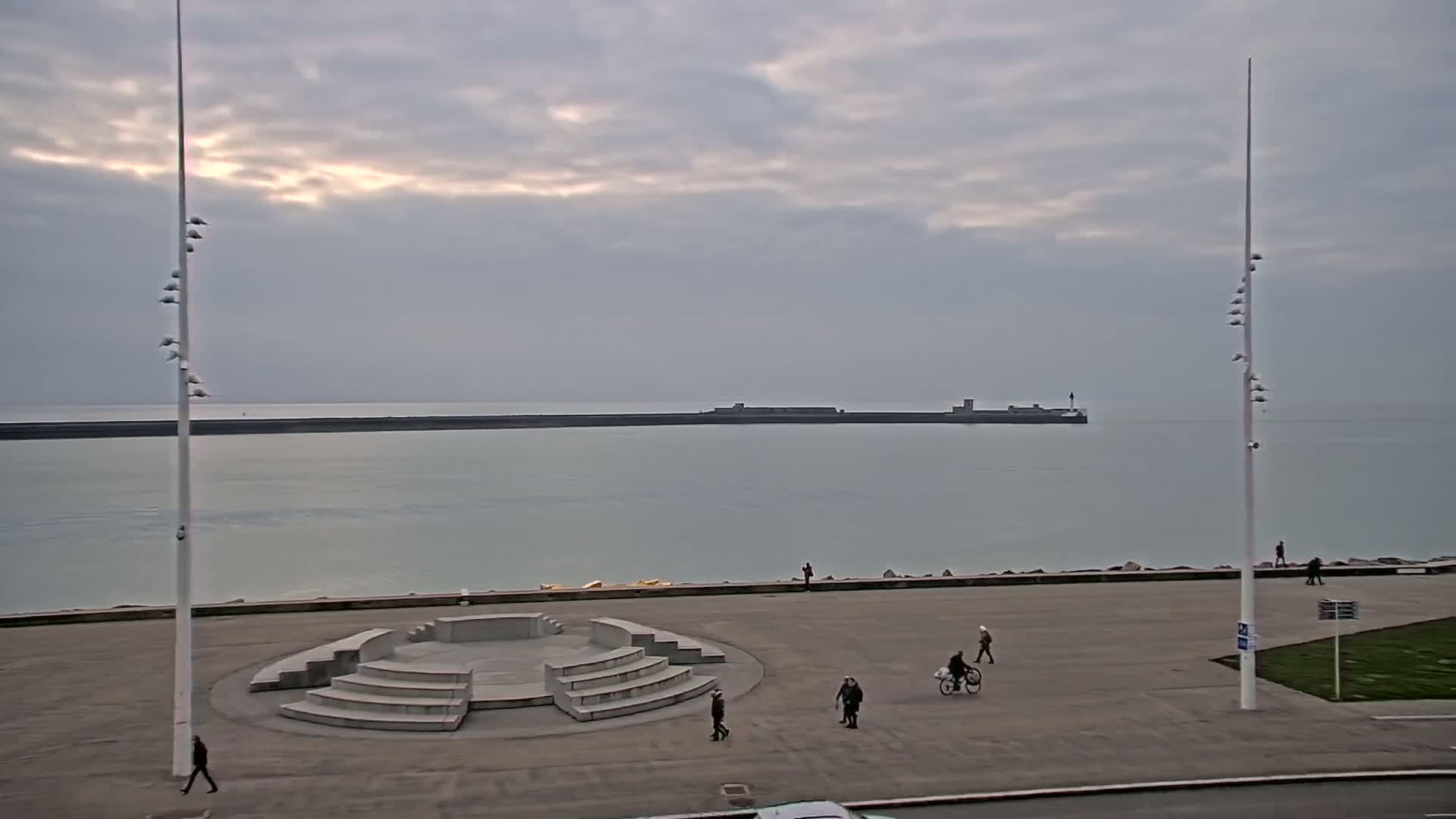 A gray, overcast day shows a calm body of water with a long breakwater in the distance, behind a paved waterfront area with a concrete amphitheater-like structure and several pedestrians.
