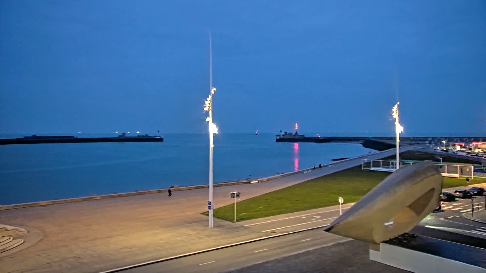 A twilight view of a calm harbor with two tall illuminated light poles in the foreground, a paved walkway, and a large abstract sculpture.