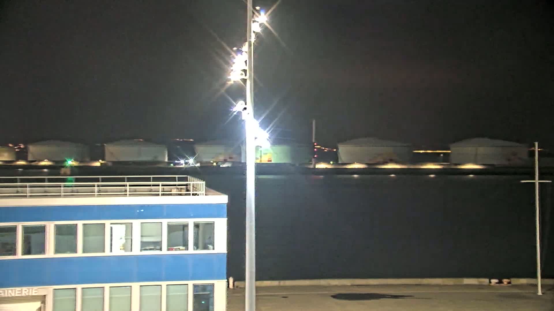 A brightly lit, blue building stands in the foreground near a body of dark water at night, with several large storage tanks illuminated across the water.
