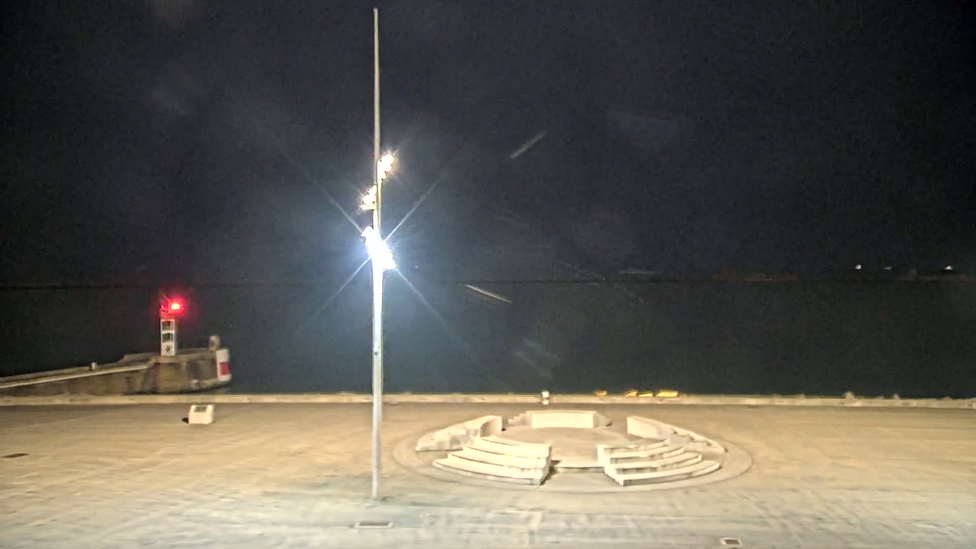 A nighttime view of a harbor with a circular stone seating area, a tall light post, and a small lighthouse in the distance.