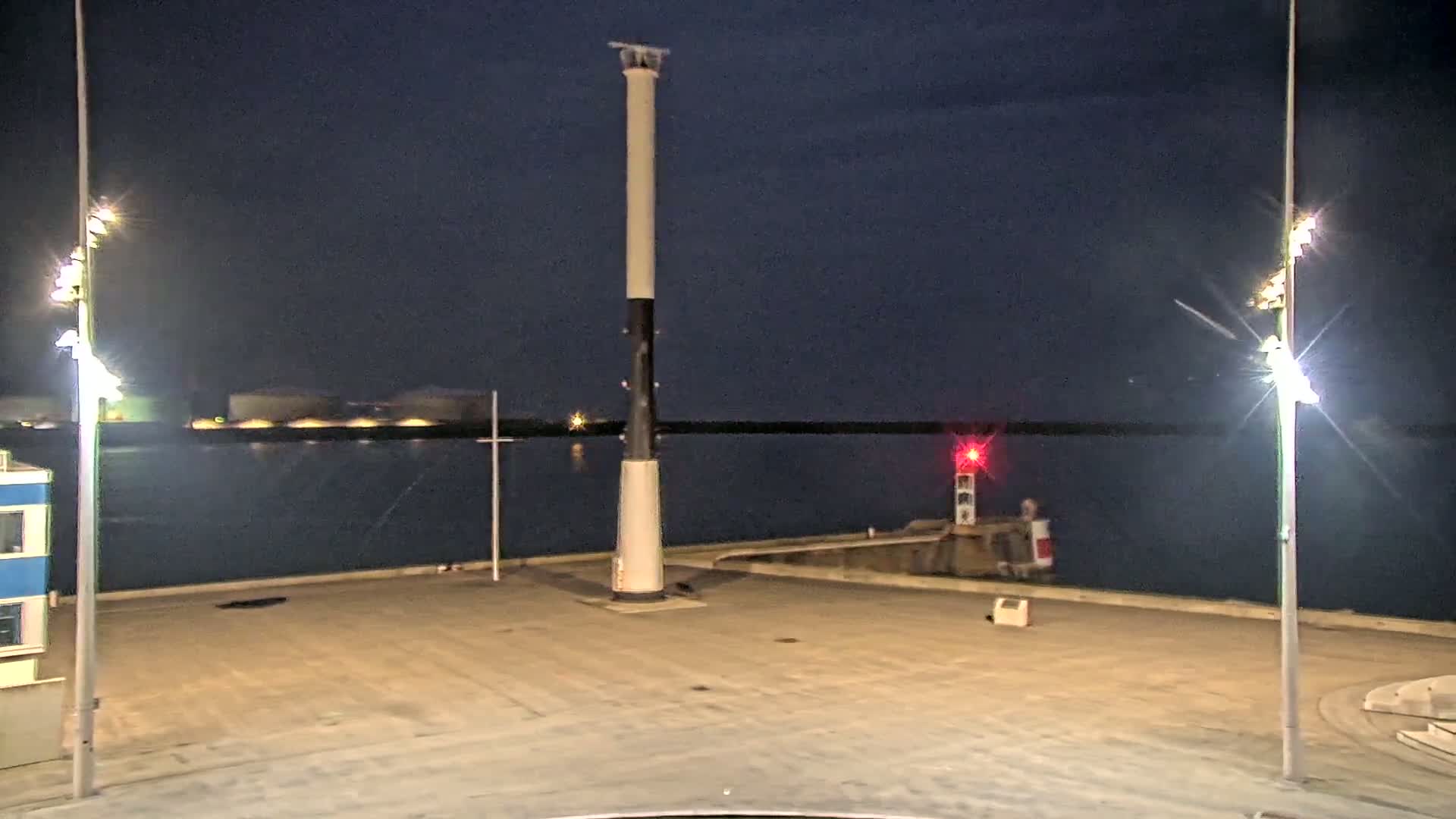 A tall, black and white lighthouse stands in a dark, empty harbor at night, illuminated by bright lights on either side.