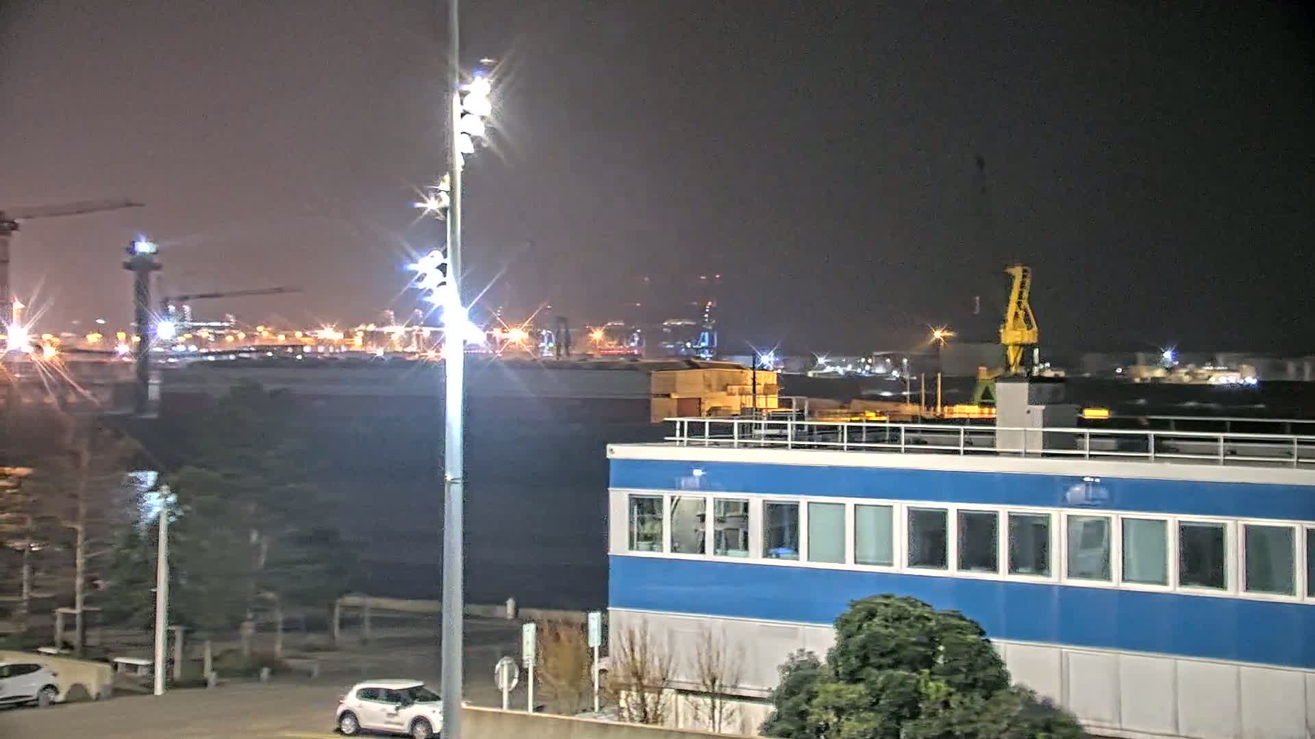 A nighttime view of a harbor with a large blue building in the foreground, a yellow crane in the middle ground, and a brightly lit cityscape in the background under a hazy sky.