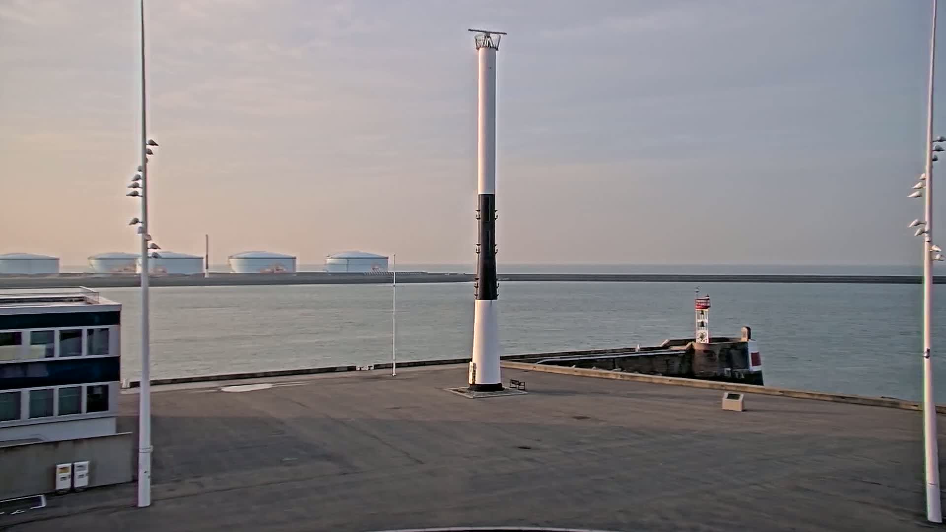 A tall, slender lighthouse stands in a harbor with calm water, several storage tanks in the distance, and a smaller lighthouse on a nearby pier under a pale sky.