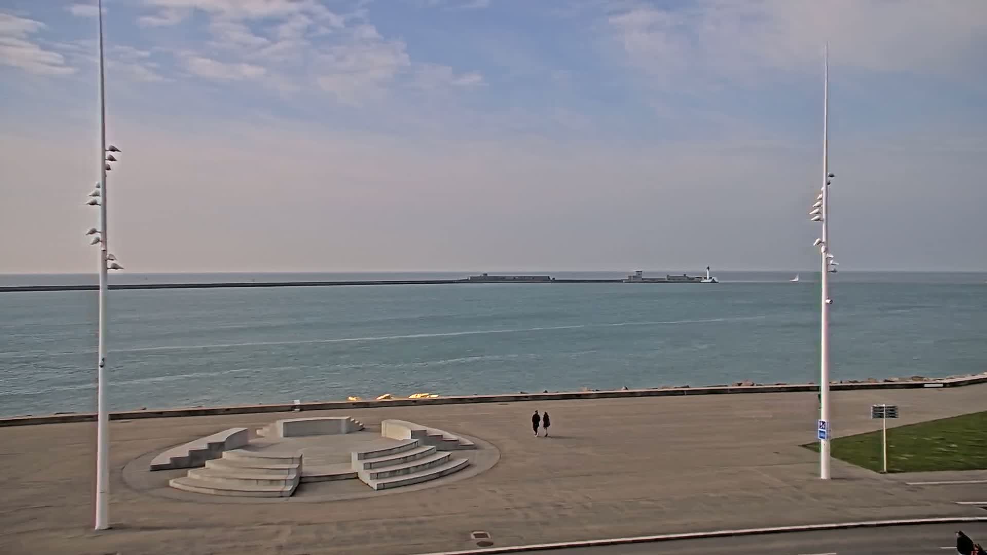 A mostly sunny day shows a couple walking on a paved plaza overlooking a calm ocean with a distant breakwater and tall, slender light poles on either side of the scene.