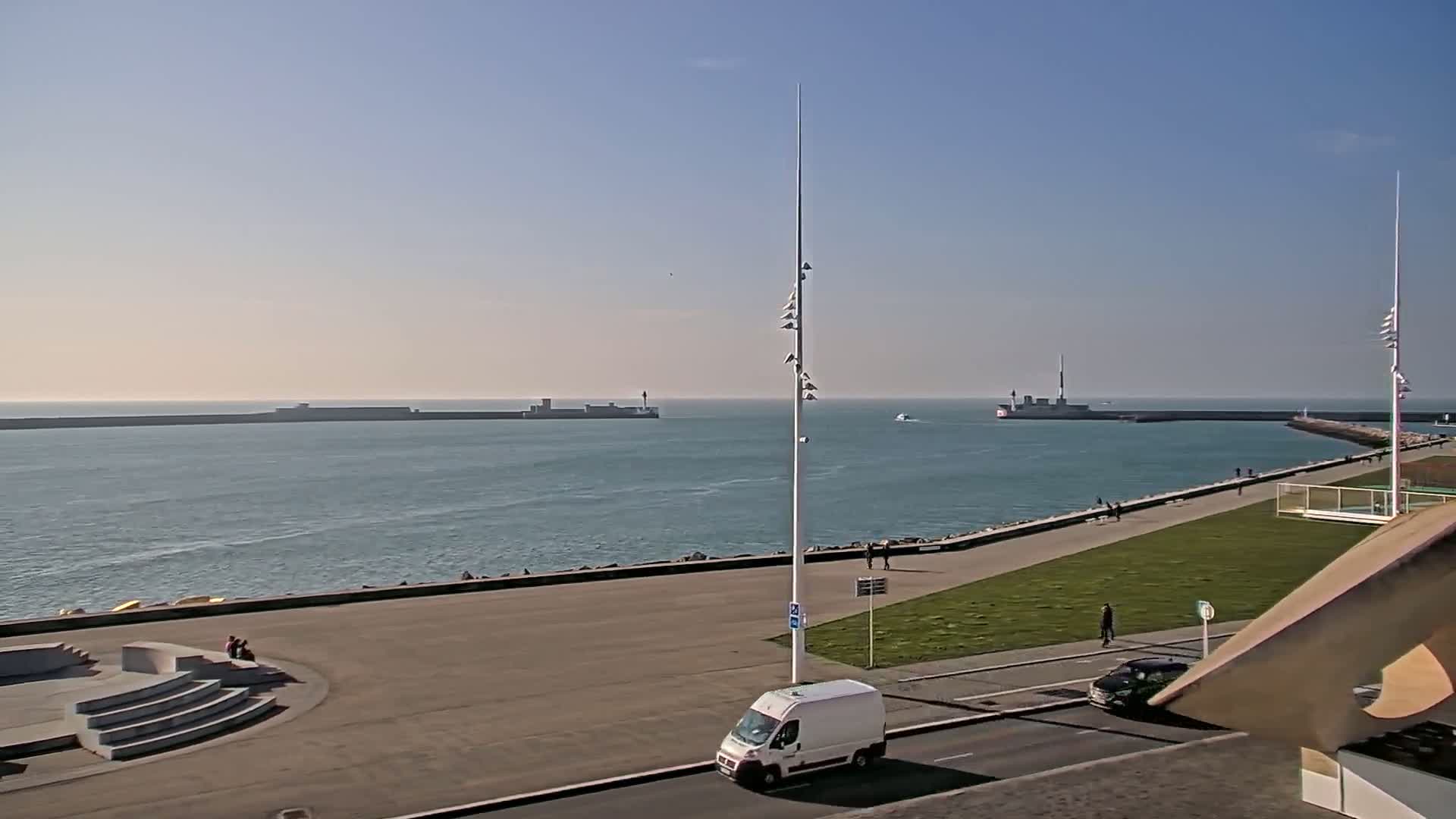 A harbor scene on a clear, sunny day shows a calm body of water, breakwaters, a paved walkway with a few pedestrians, and a white van parked on a road.