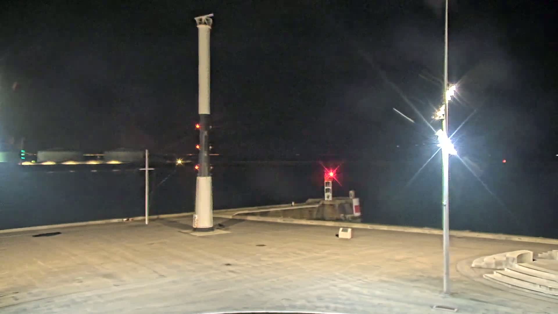 A tall, slender white and black column stands in a dark, empty harbor area at night, next to a smaller lighthouse and under bright lights.