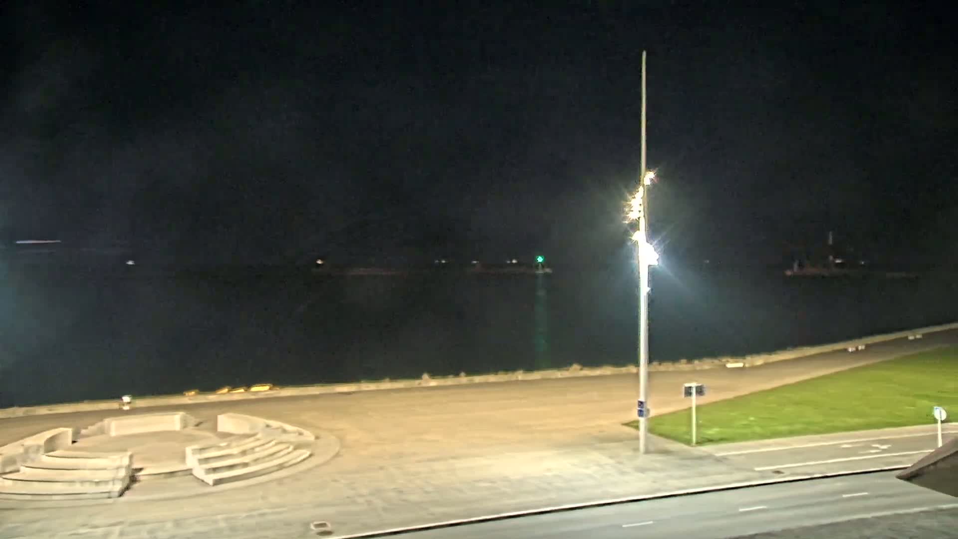 A nighttime view of a calm body of water, a paved waterfront area with a circular seating structure, a grassy area, and a roadway.