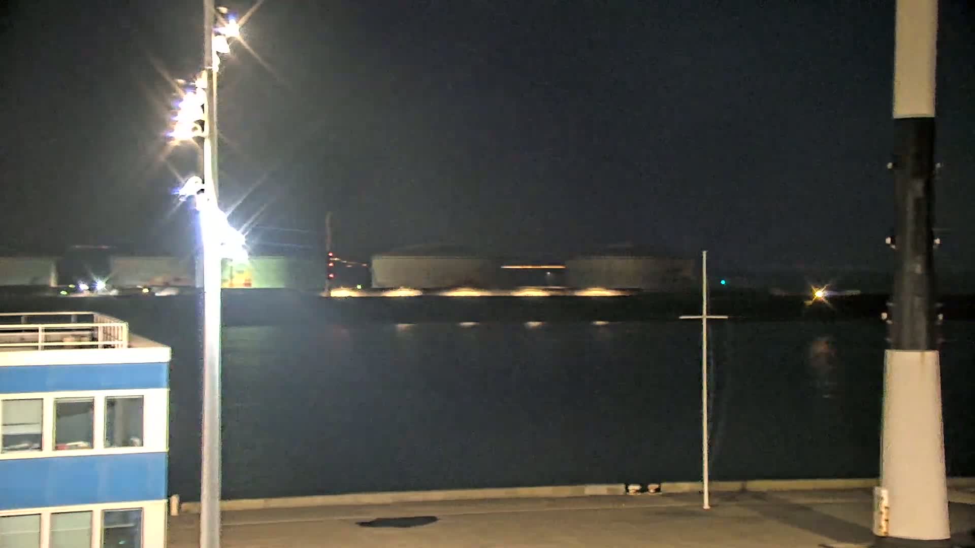 A nighttime view of a body of dark water, with a building and tall light post in the foreground, and industrial buildings across the water.