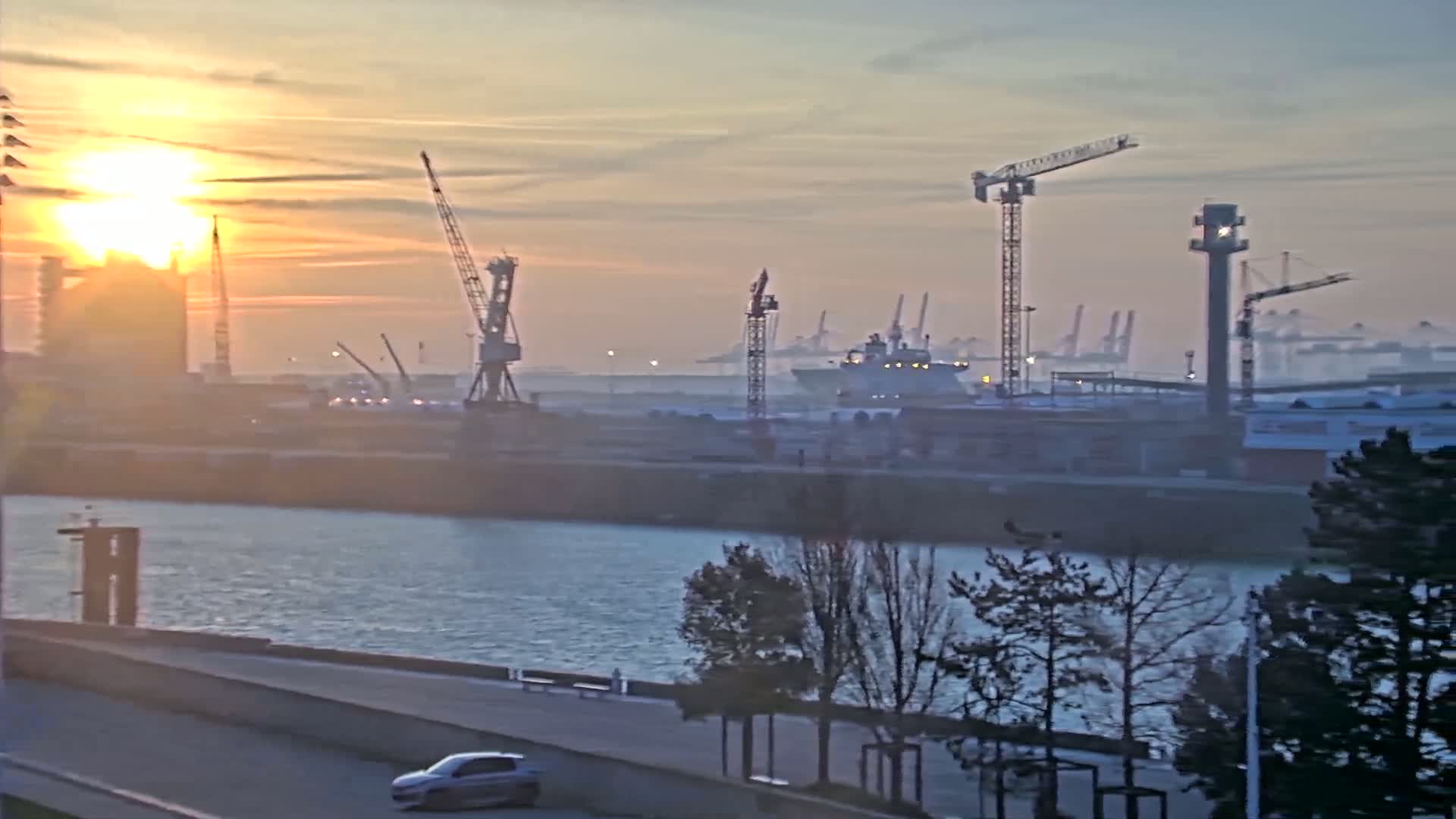 A hazy sunset illuminates a harbor scene with numerous cranes, ships, and a car driving along a waterfront road.