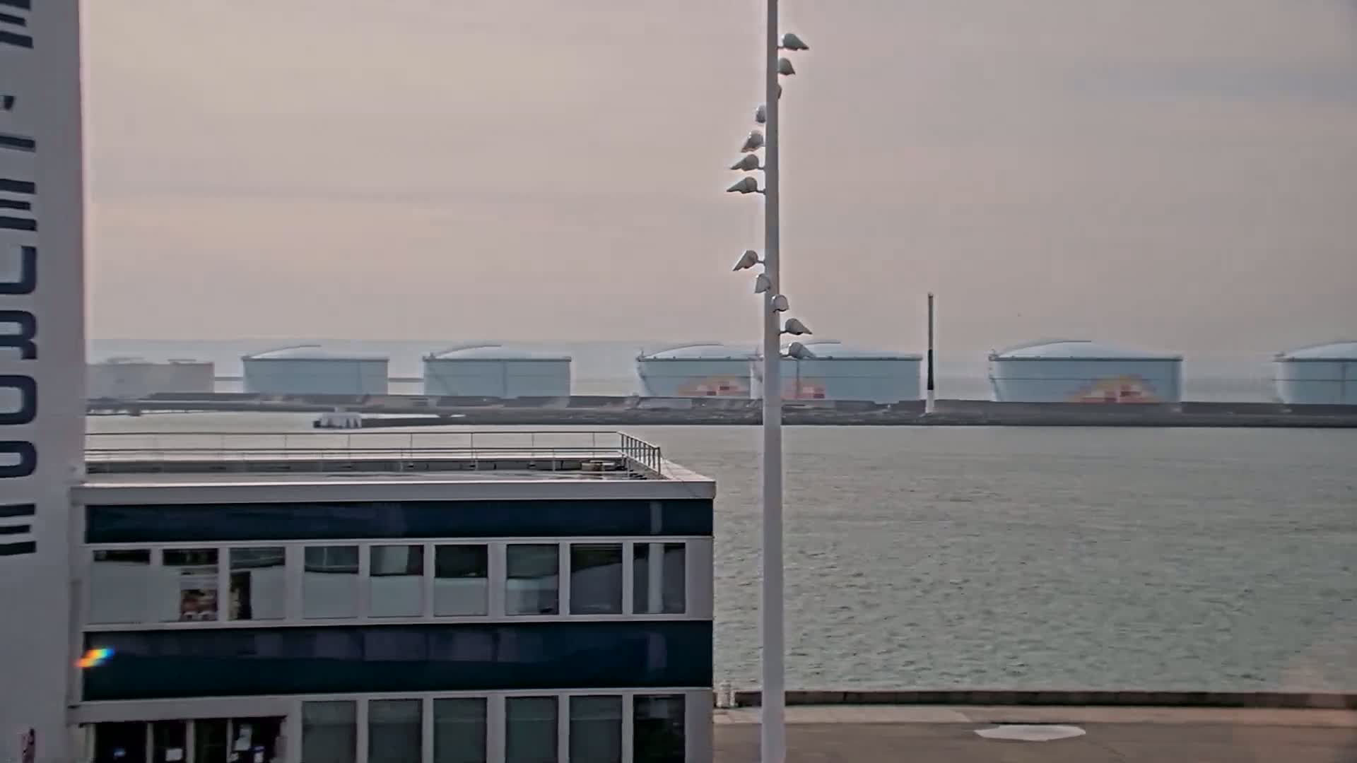 A multi-story building with many windows is seen in the foreground, with a row of large storage tanks across a body of water in the background on an overcast day.