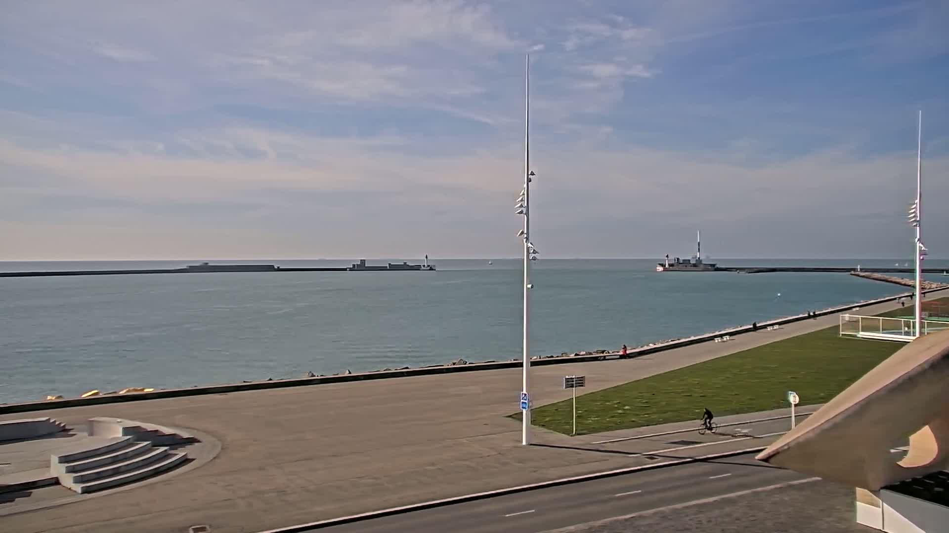 A calm, partly cloudy day at a harbor, showing a paved waterfront area with a grassy lawn, a bicycle rider, and a ship in the distance.
