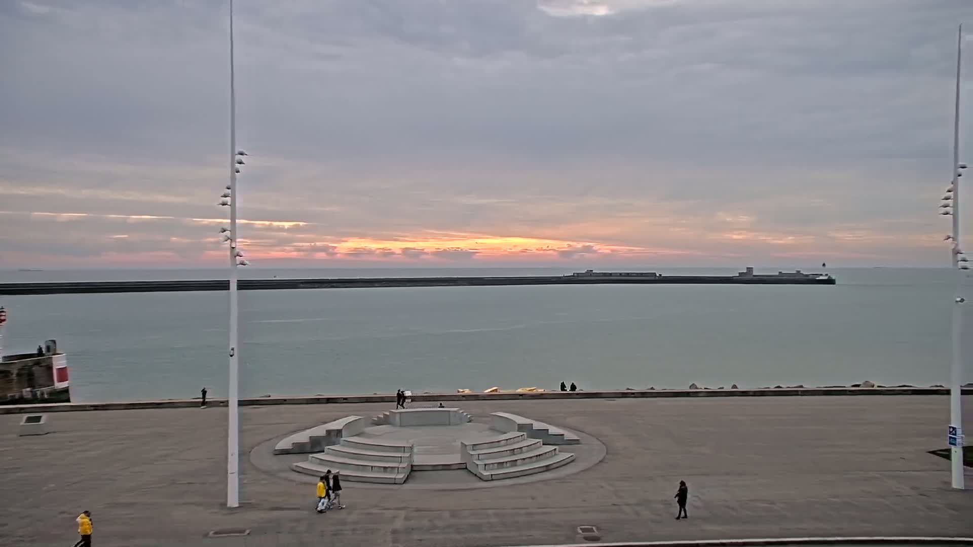 A calm sea under an overcast sky with a pale orange sunset, viewed from a paved waterfront area featuring a circular stone amphitheater and several people scattered about.