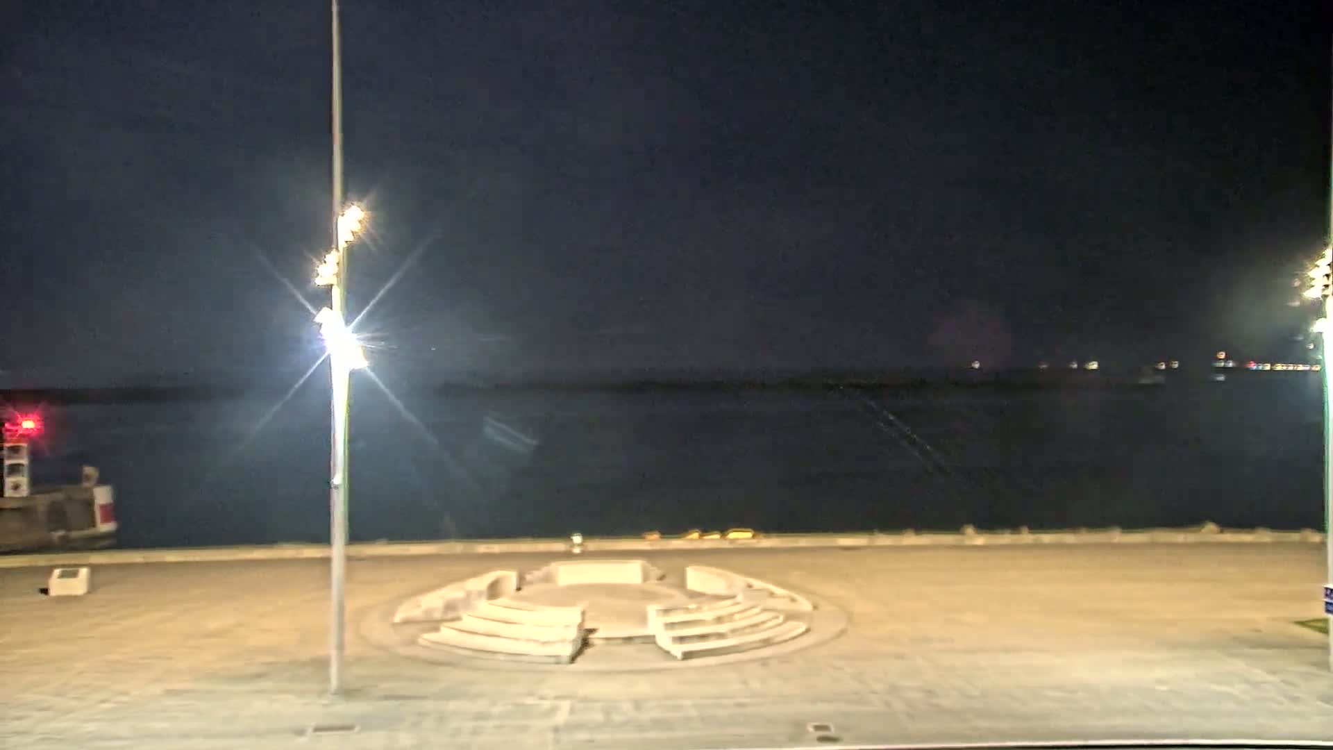 At night, a circular amphitheater-like structure sits in a paved plaza overlooking a calm body of water under a dark sky.