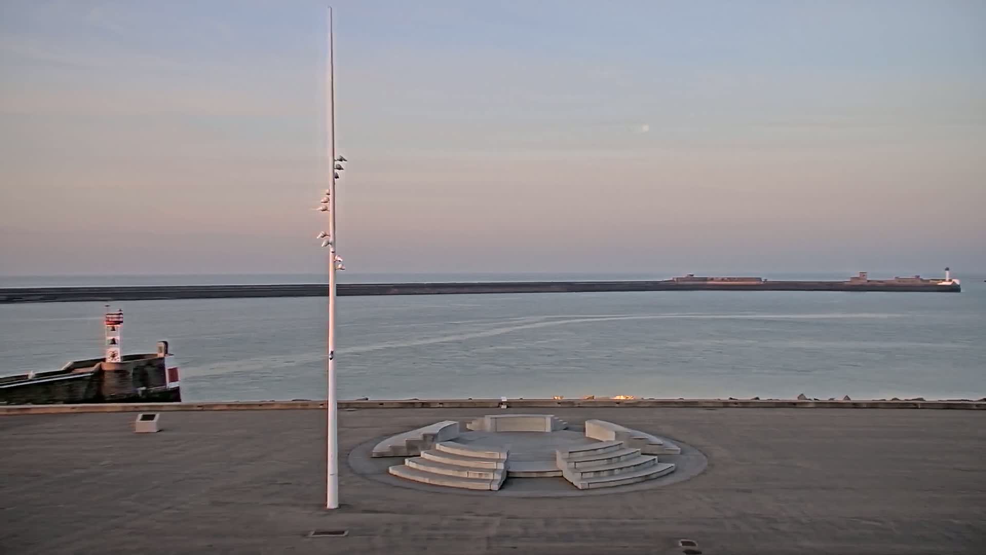 A tall flagpole stands in a paved area overlooking a calm sea with a breakwater and lighthouse in the distance under a pale sky.