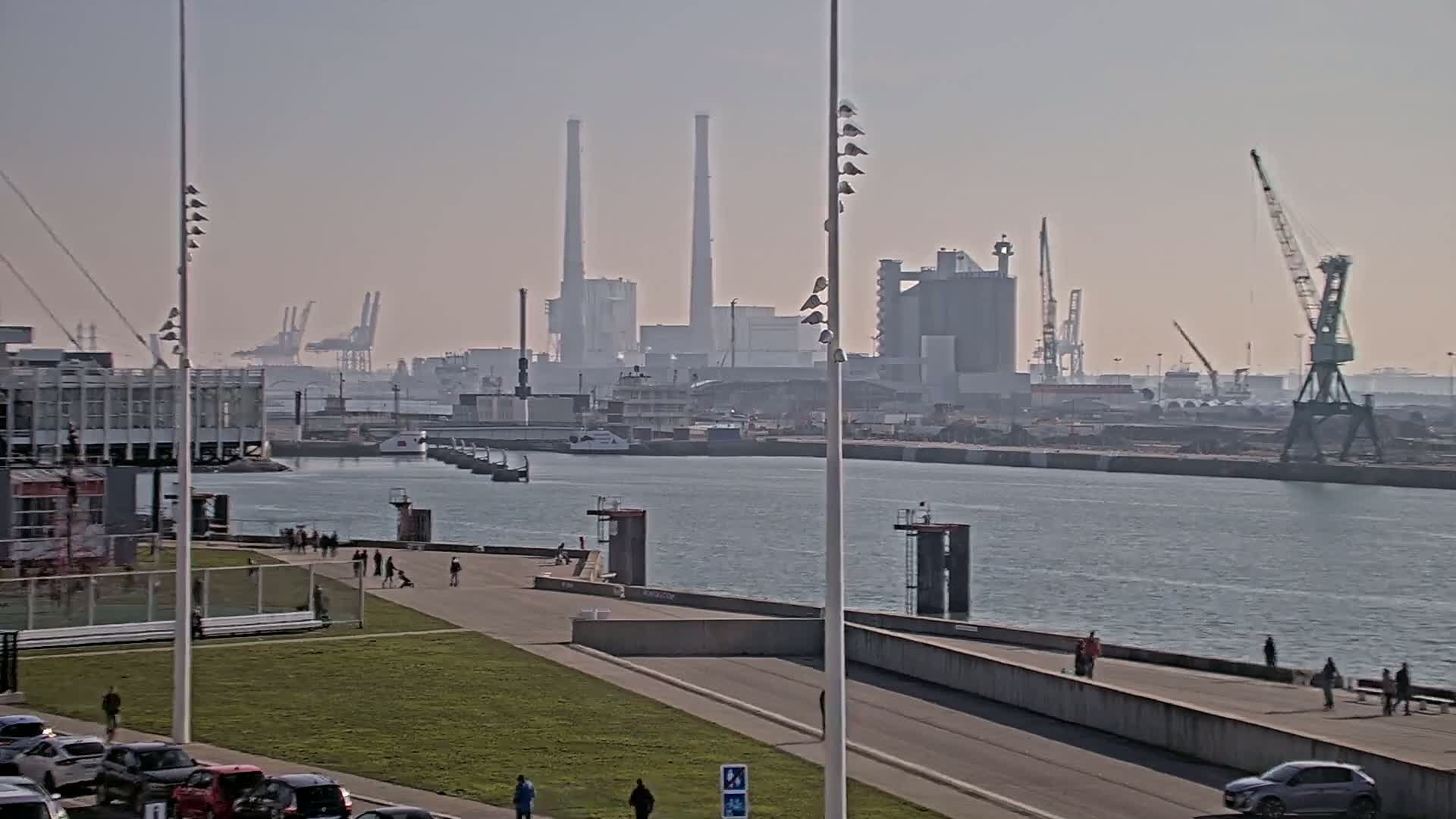 A hazy day reveals a harbor scene with industrial buildings, cranes, and a calm body of water, where people stroll along a paved waterfront path.