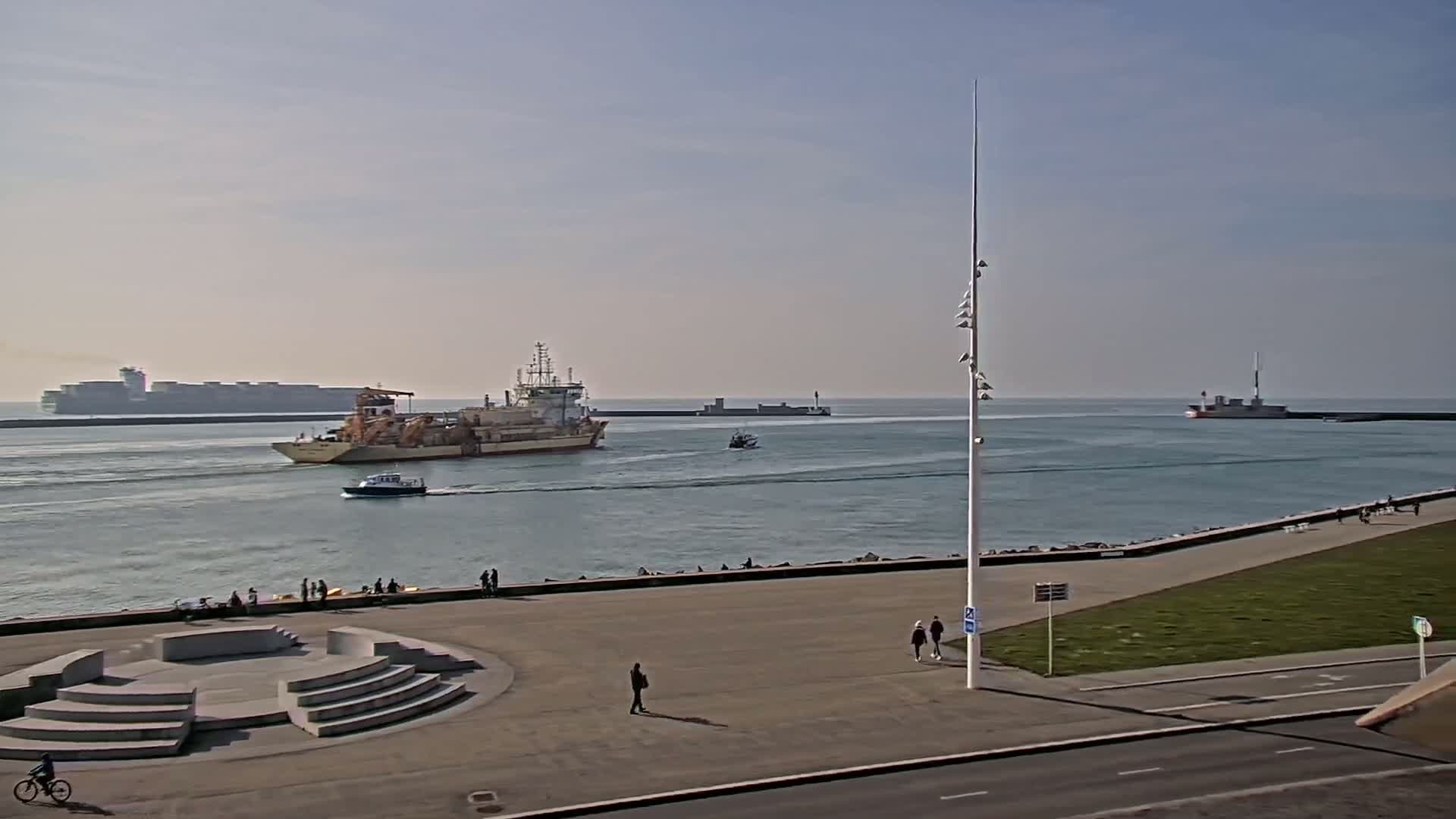 A harbor scene on a calm, sunny day shows several ships including a large cargo ship in the distance, a dredging vessel, and a small boat; a paved area with a few people and a bicycle is in the foreground.