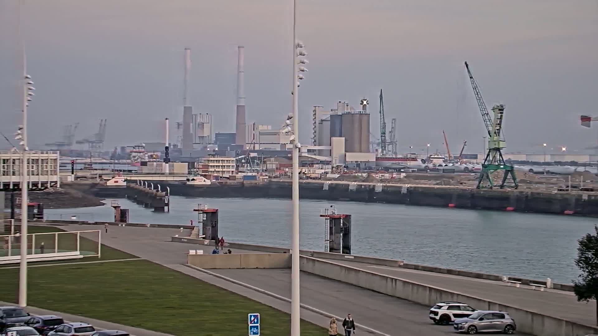 A waterfront area with a paved walkway and grassy areas overlooks a harbor with industrial buildings and cranes under an overcast sky.