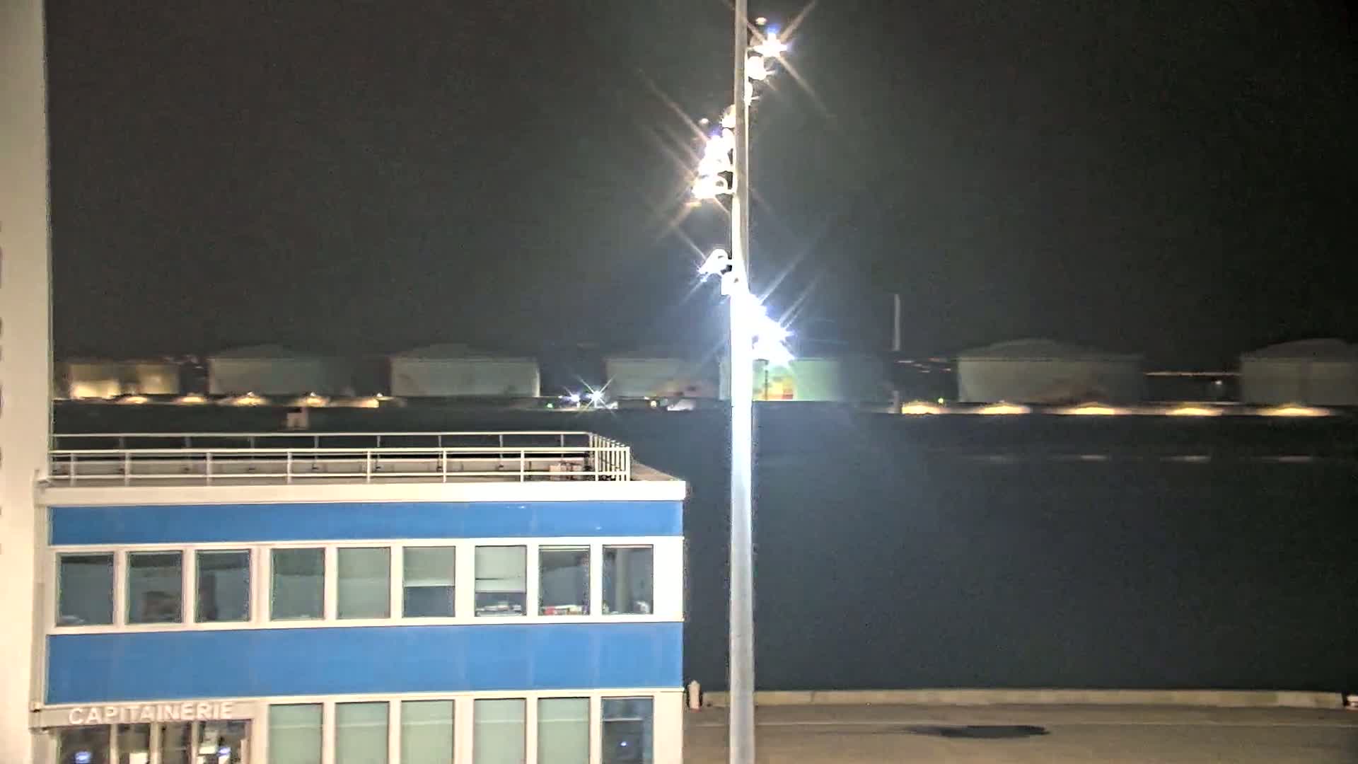 At night, a brightly lit tall pole stands beside a blue building next to a dark body of water with illuminated structures in the distance.