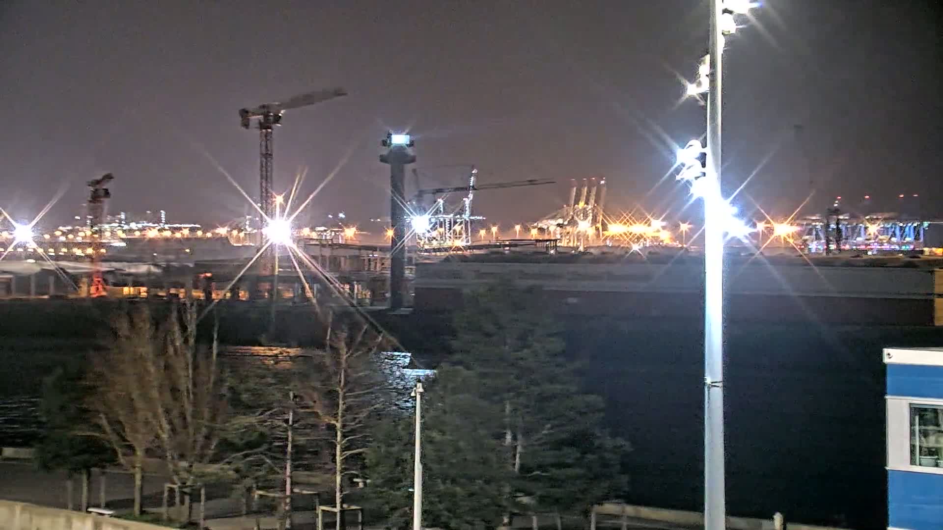 A nighttime view of a harbor with brightly lit cranes and shipping containers, viewed from across a body of water with trees in the foreground.